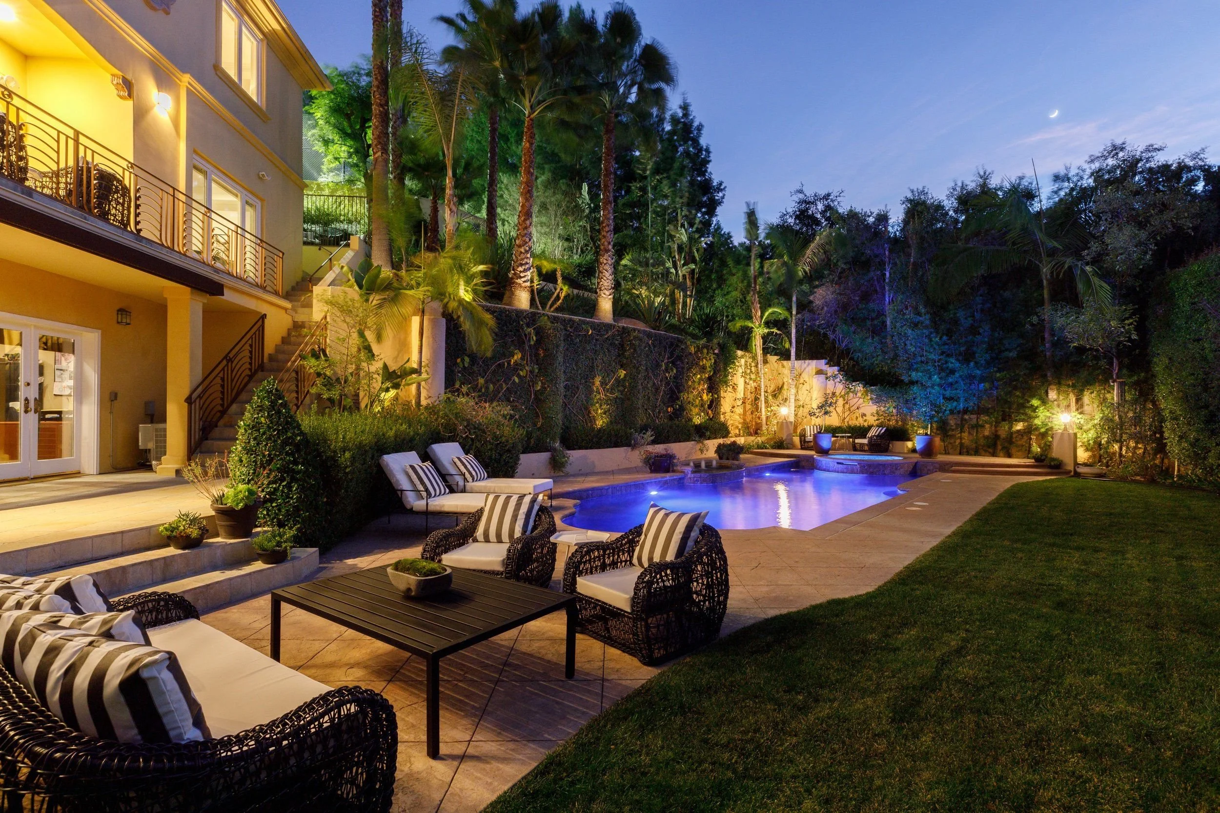 A backyard pool area at night with outdoor furniture, lush trees, and a tiled patio, illuminated by lights, with a house featuring a staircase and balconies on the left.