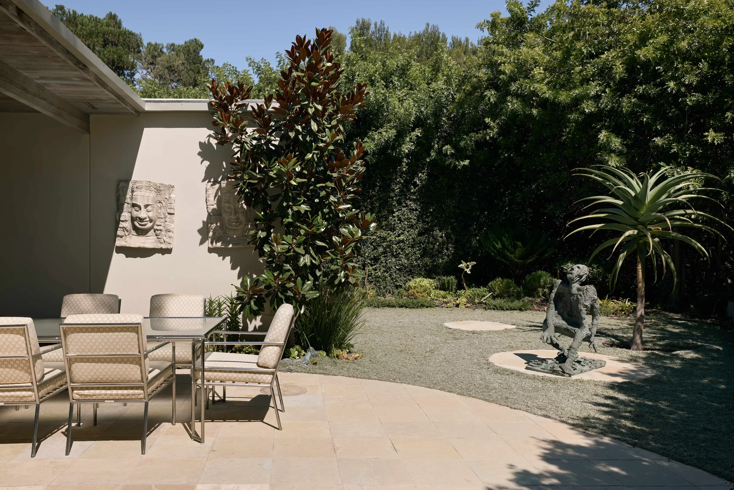 An outdoor patio with a dining table and chairs next to a white wall decorated with two stone carvings of smiling faces. The patio is adjacent to a garden with various trees and plants, including a large leafy shrub and a statue of a seated animal.