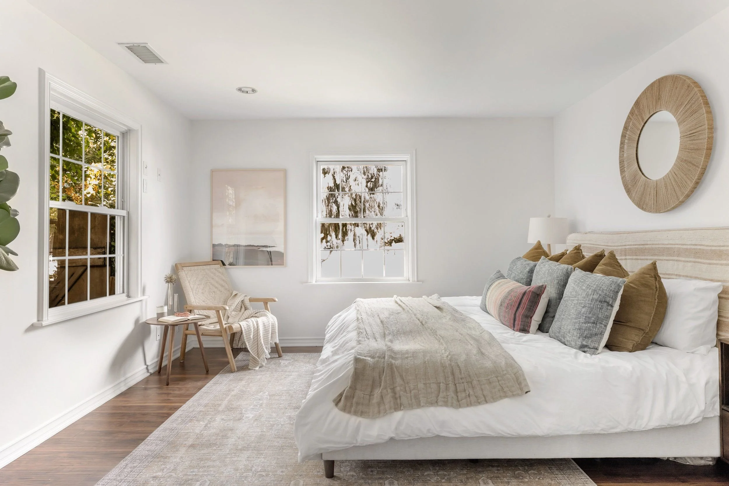 A cozy bedroom with white walls, large windows showing trees outside, and a wooden floor. There is a bed with a beige headboard, gray and beige pillows, and a light-colored blanket. To the left, there is a small wooden chair with a blanket draped ove
