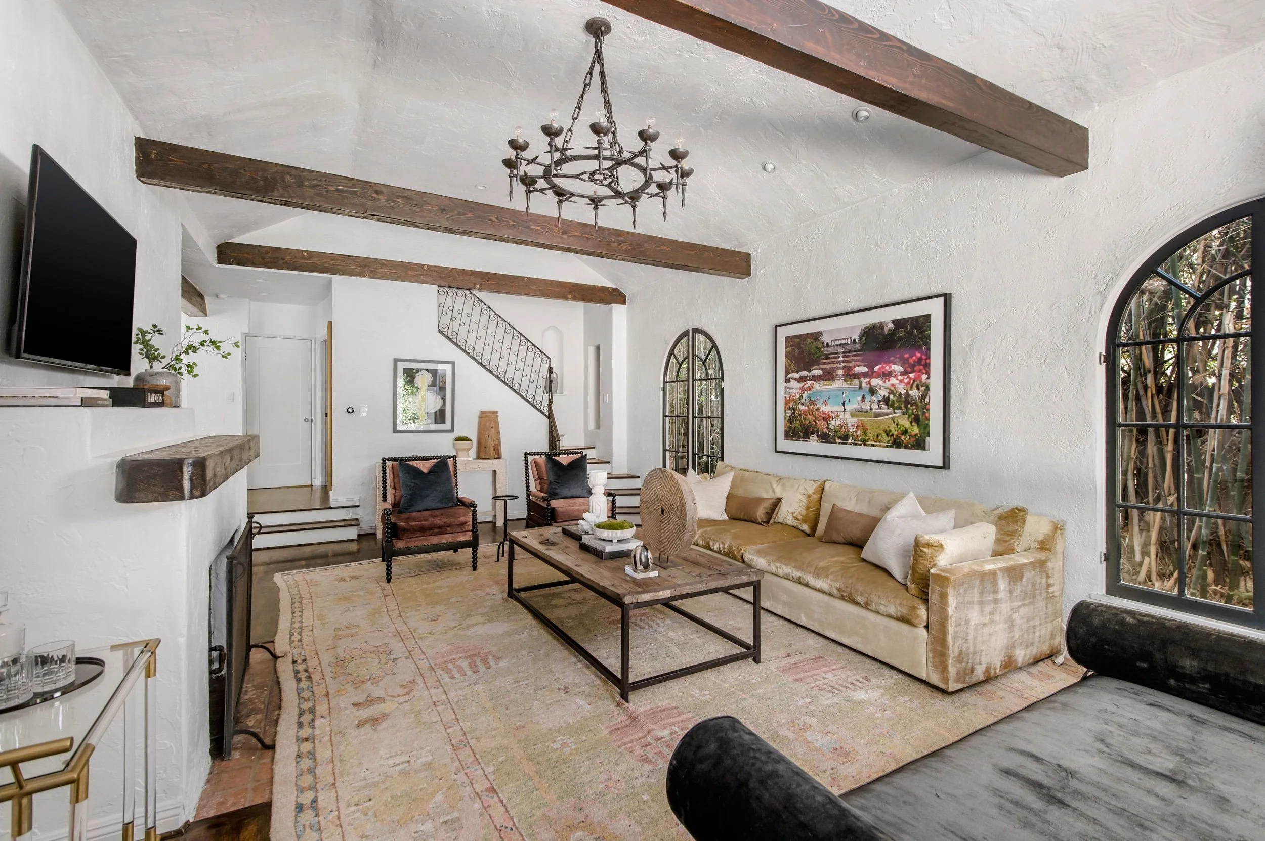 A living room with white textured walls and ceiling featuring exposed dark wooden beams, a large window with an arched black frame, a beige sofa with pillows, two dark armchairs, a rustic wooden coffee table, a TV, and decorative artwork and plants.