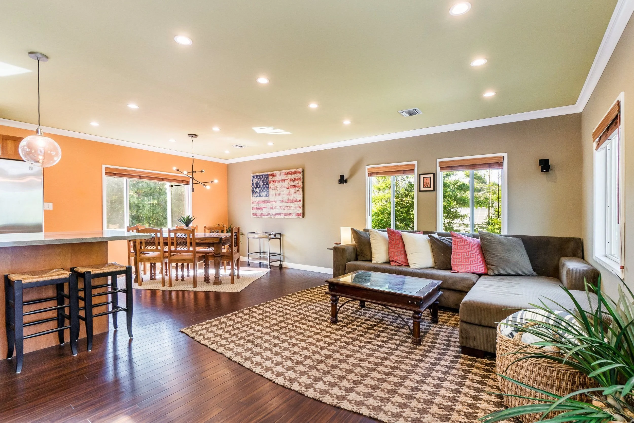 Living room with hardwood floors, beige walls, large windows with blinds, a gray sofa with pillows, a wooden coffee table, a patterned area rug, and a dining area with a wooden table and chairs. Recessed lighting and a modern chandelier.