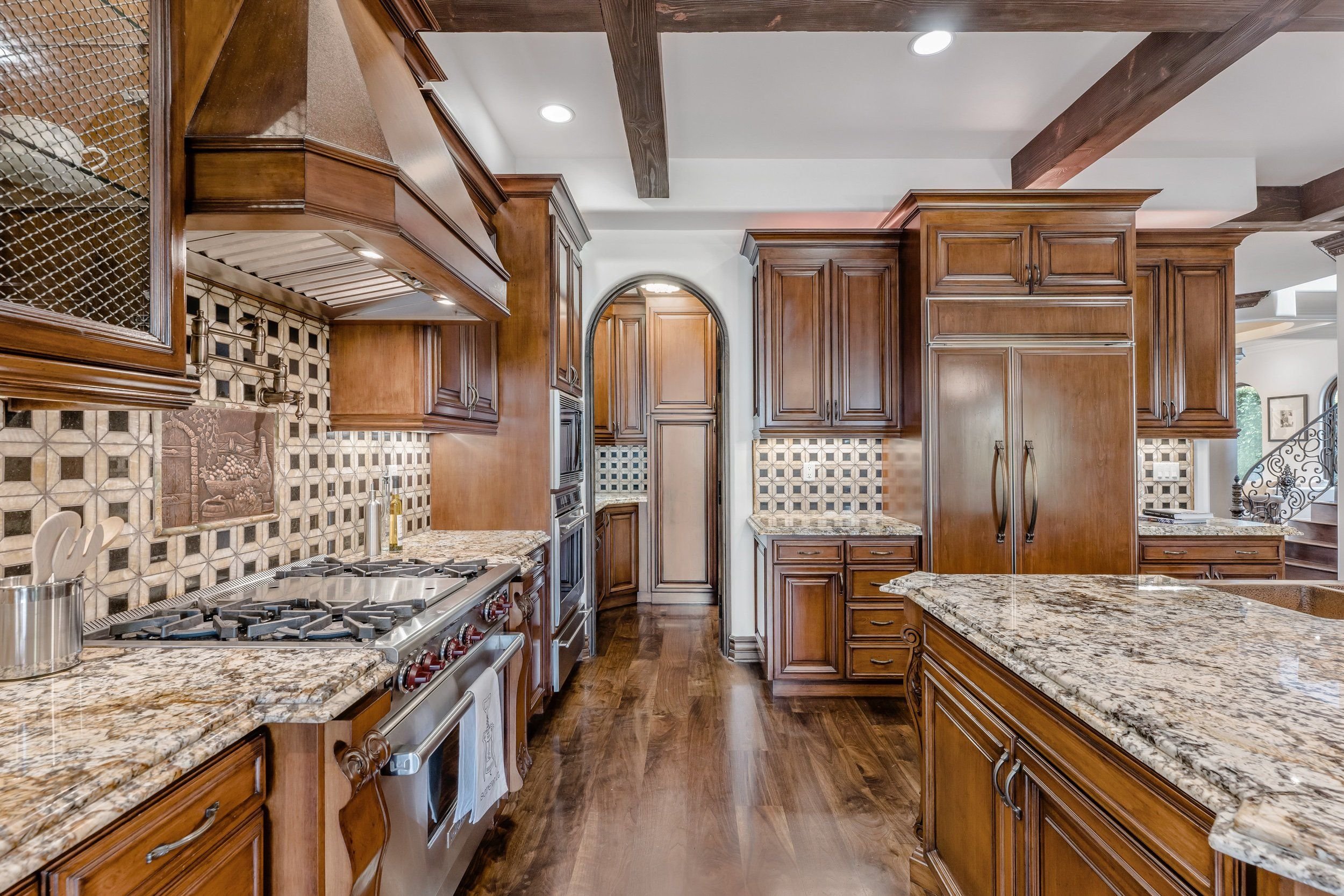 A spacious kitchen with wooden cabinetry, granite countertops, and a backsplash with artistic tile patterns. It features a stainless steel stove and oven, a double sink, and a large kitchen island. The ceiling has recessed lighting and wooden beams.