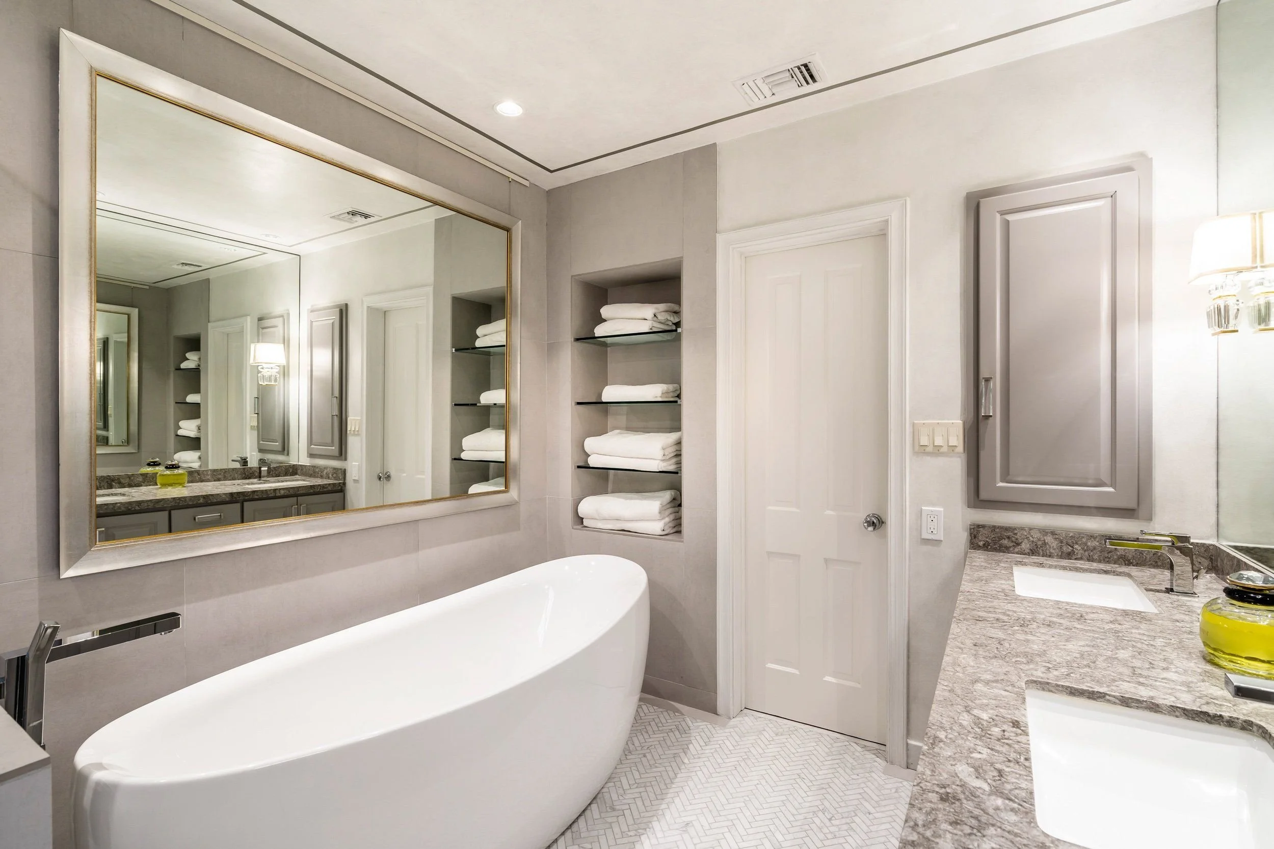 Modern bathroom with a large mirror, a freestanding bathtub, an open shelf with folded towels, and a vanity with a granite countertop.