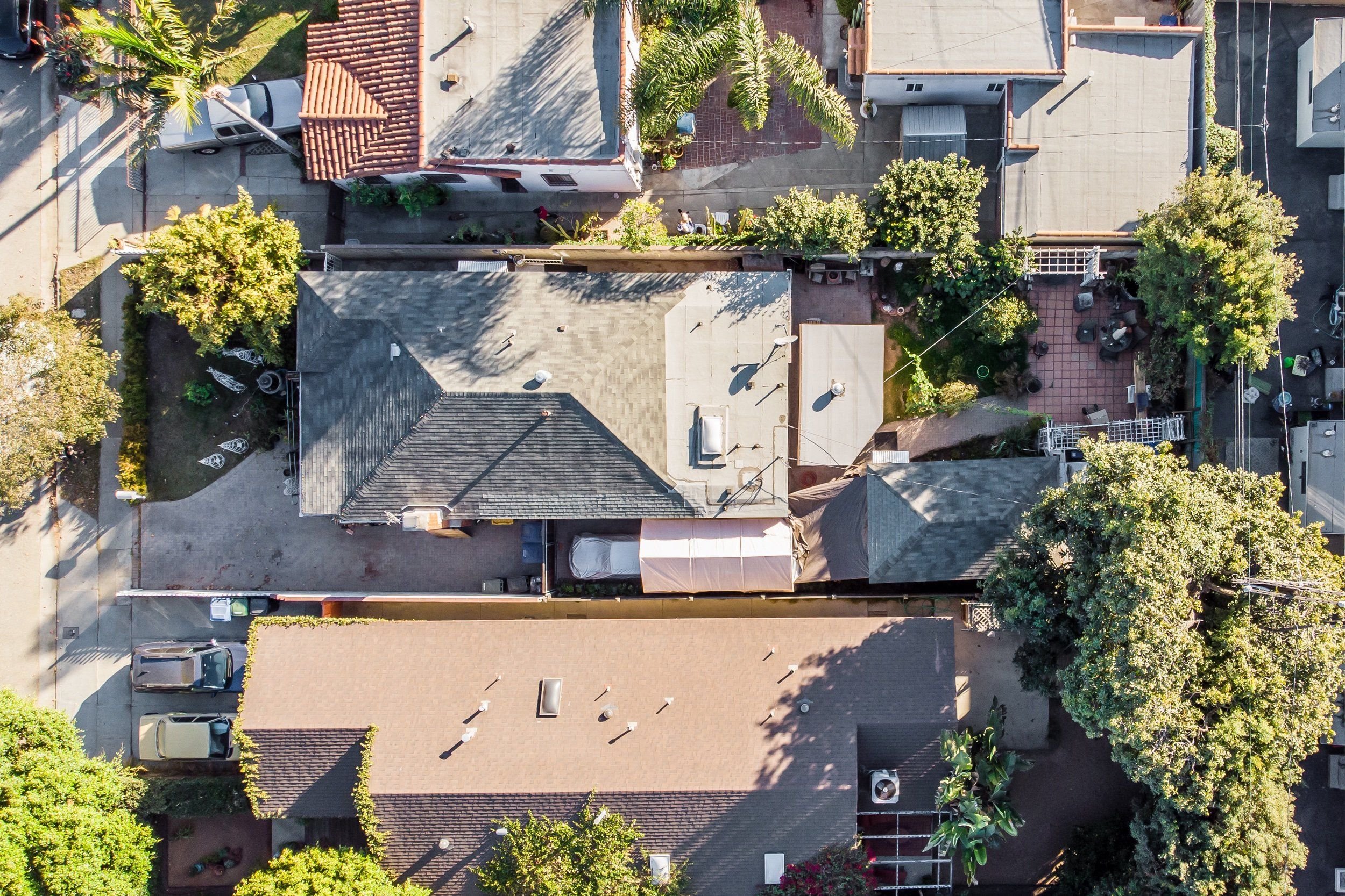 An aerial view of residential houses with rooftops, trees, cars parked along the streets, and a small backyard patio.