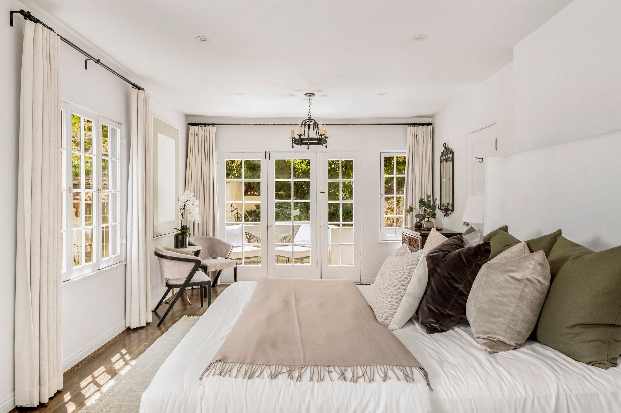 Bright living room with large windows, white curtains, and a chandelier. There are a beige sofa with multiple pillows and some chairs near the windows, with potted plants and outdoor greenery visible outside.