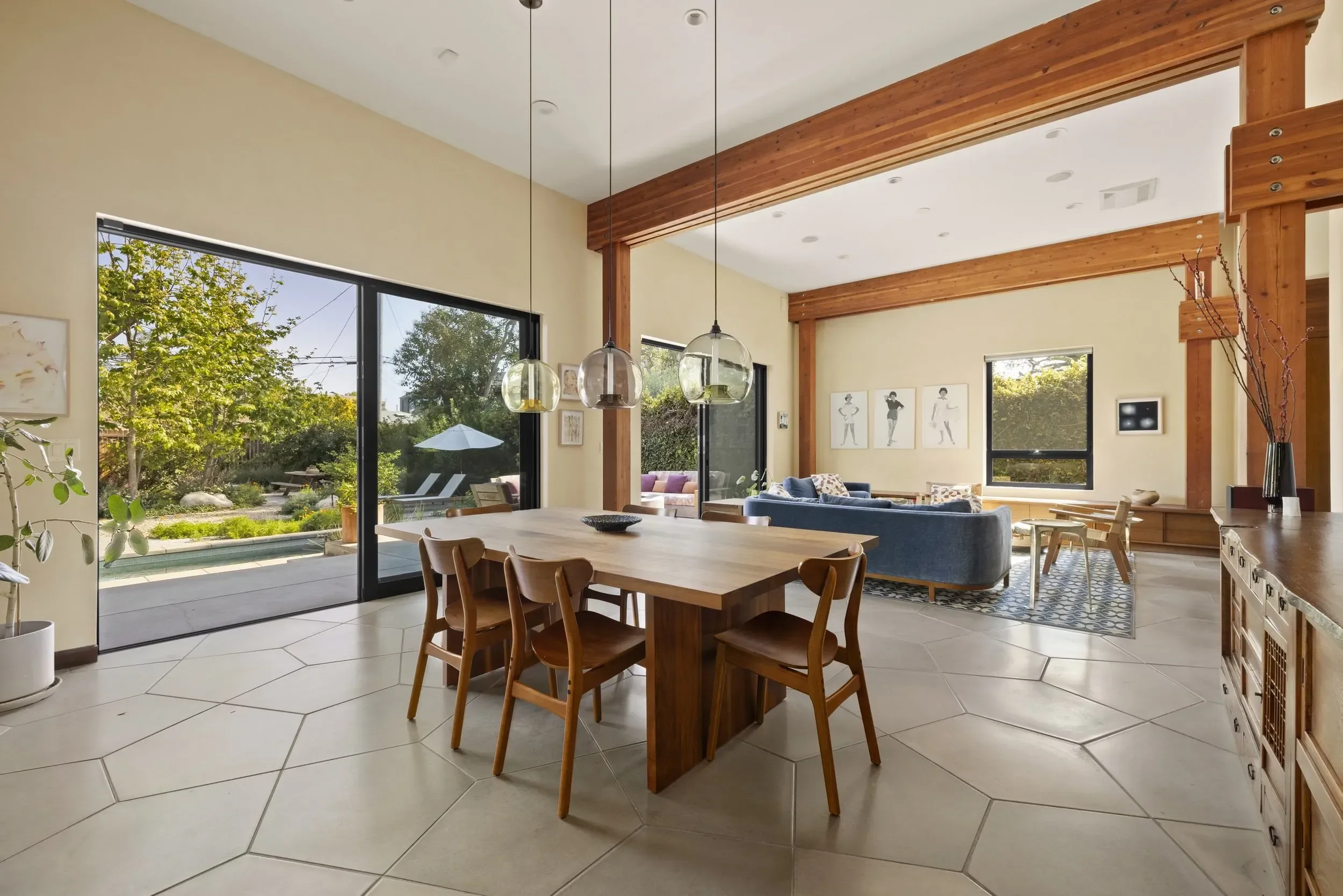 A spacious living room with large glass sliding doors leading to an outdoor patio with seating and umbrella. The room features high ceilings with wooden beams, beige walls, and tiled flooring. There are modern light fixtures hanging above a wooden di