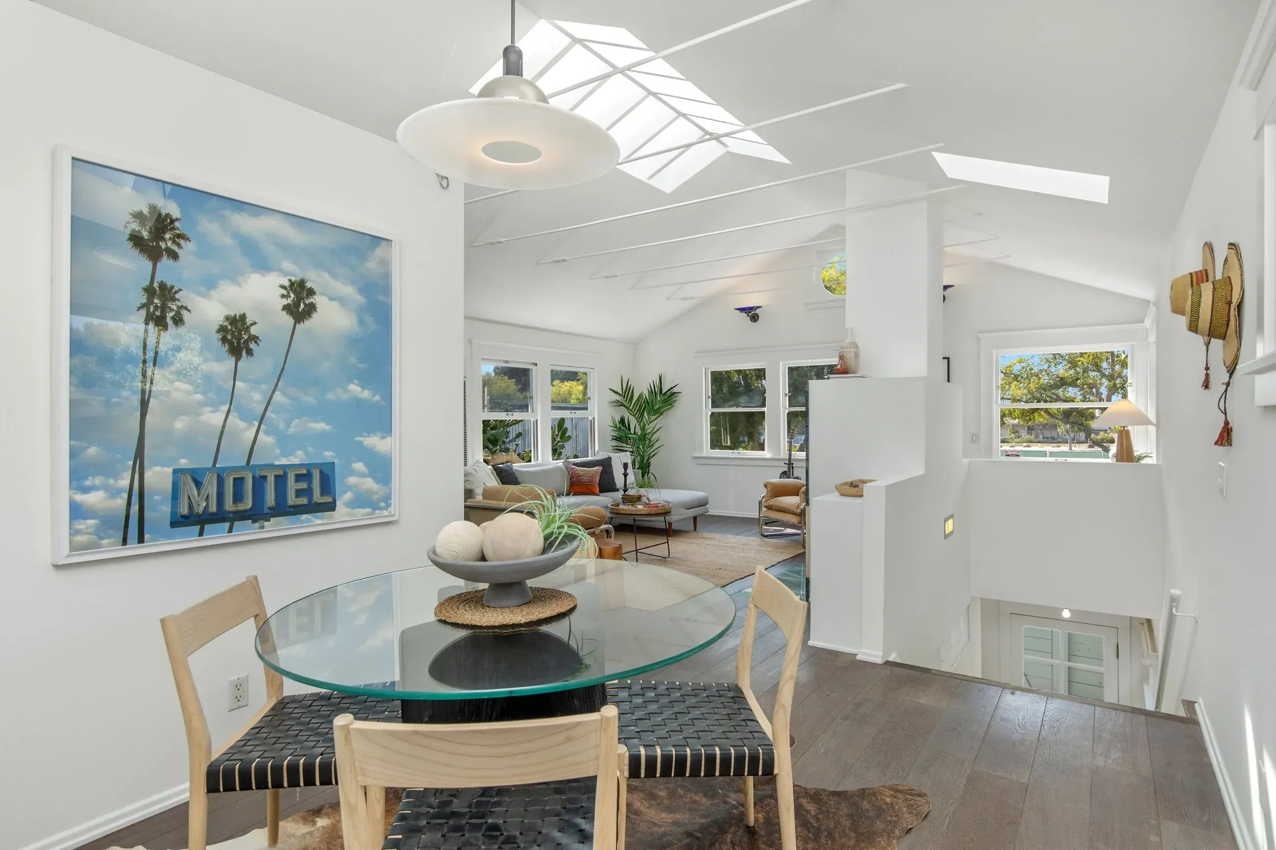 Interior of a bright, modern living room with white walls, large windows, and skylights. It features a dining area with a glass table and four light wooden chairs. The living area has a plush sectional sofa with colorful pillows, indoor plants, and a