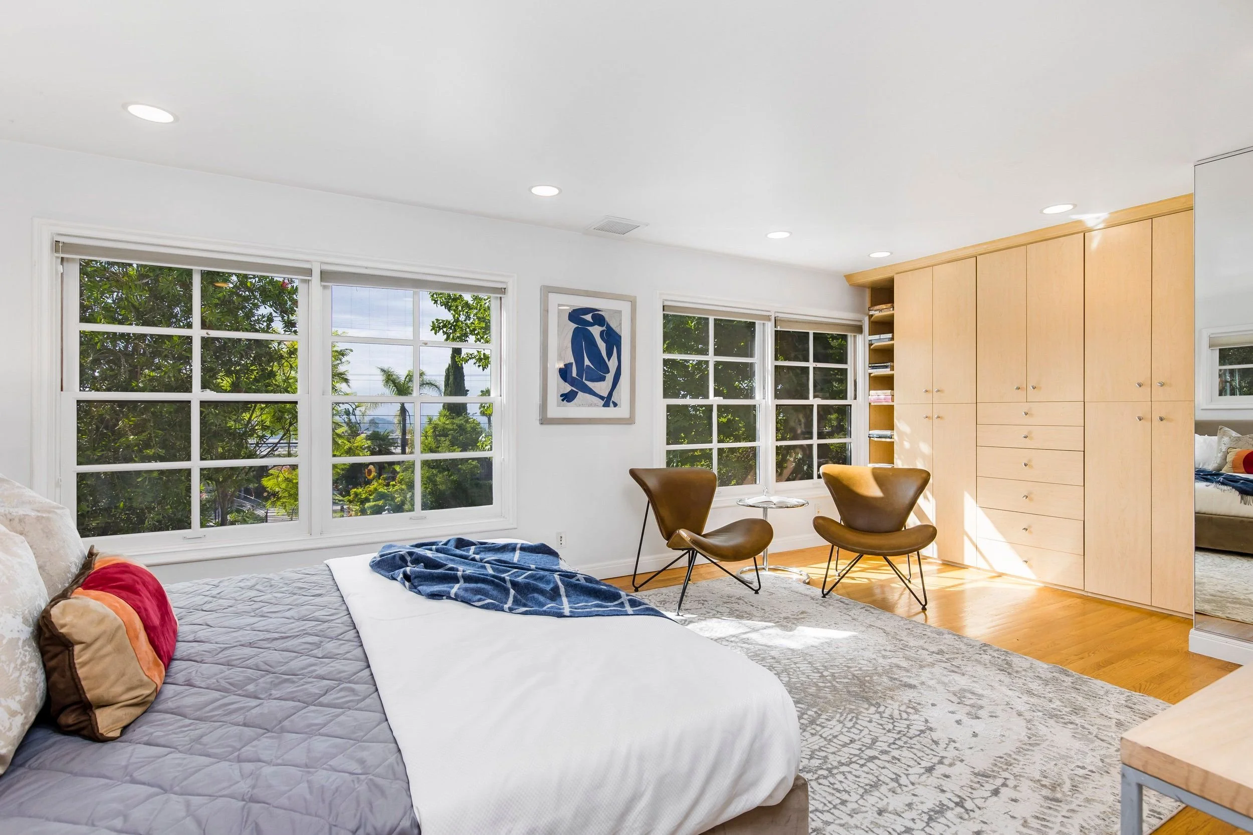 Bright living room with large windows showing trees outside, featuring a white couch with colorful pillows, two brown chairs, a light wooden built-in cabinet, and a framed abstract artwork on the wall.