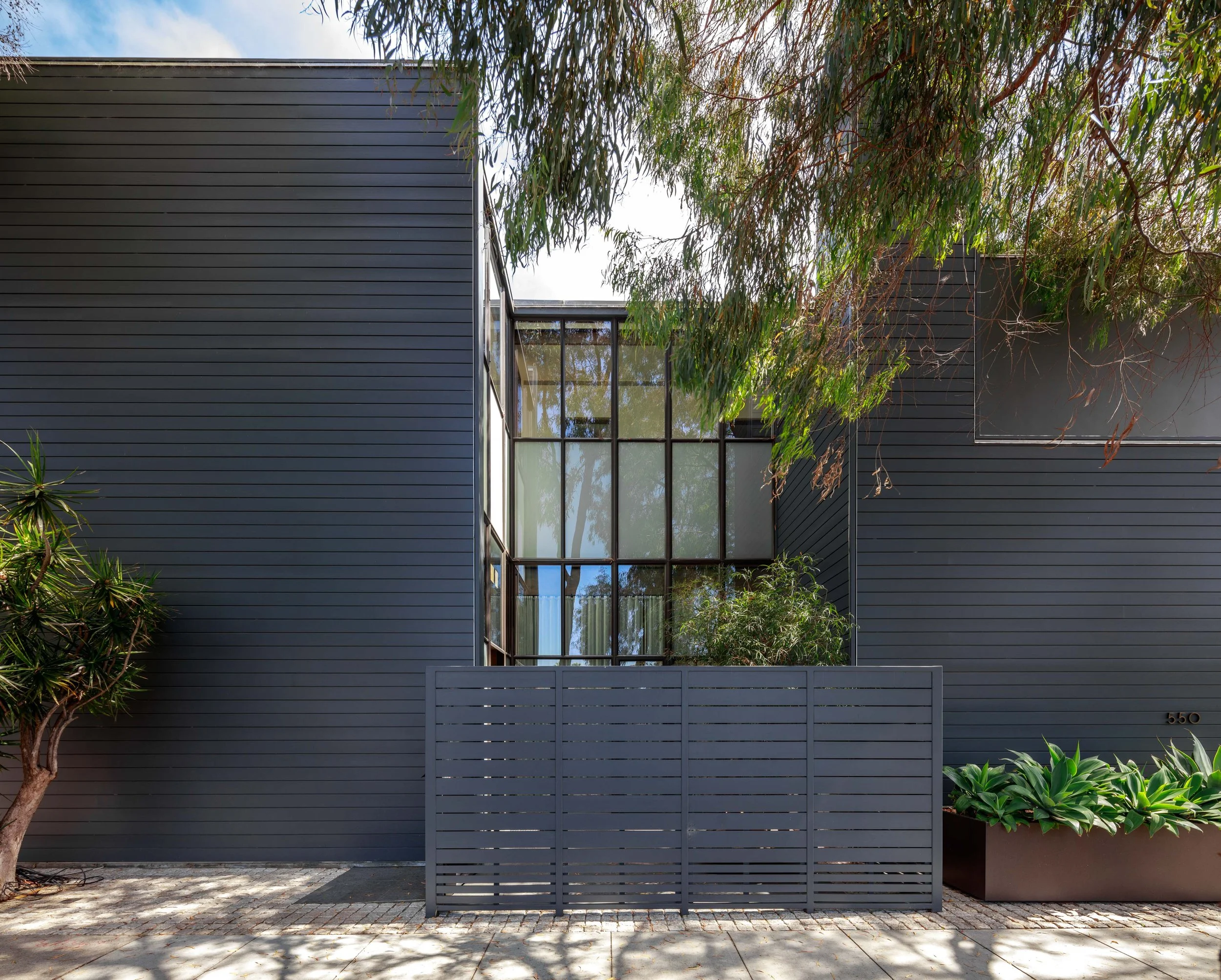 A modern building with dark gray horizontal siding, large glass windows, and trees surrounding it. The lower part shows a paved area with some shadows from the trees.
