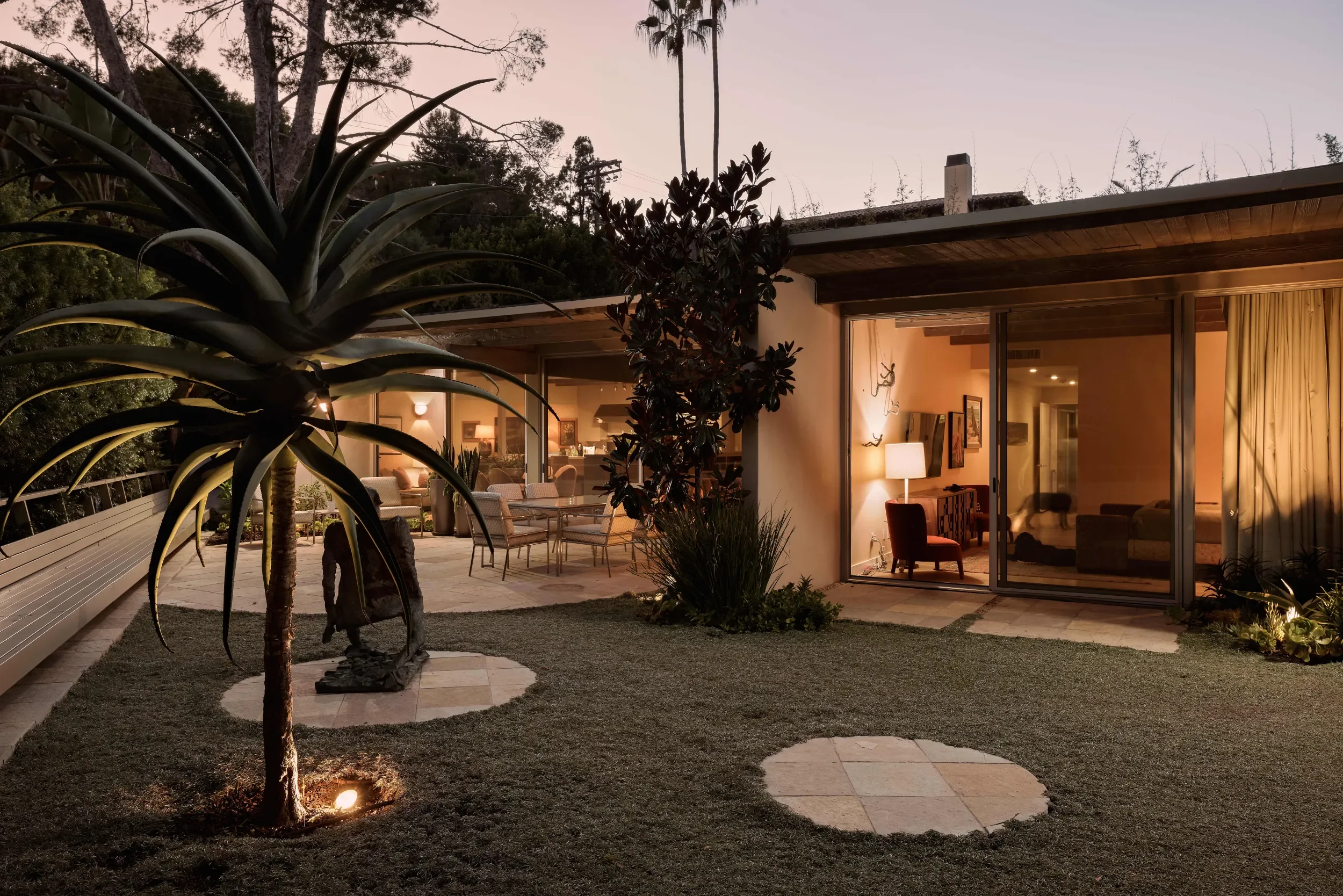 Nighttime view of a patio with outdoor furniture, large palm-like plants, and indoor living space visible through sliding glass doors. The area is illuminated by warm lighting with trees and sky in the background.