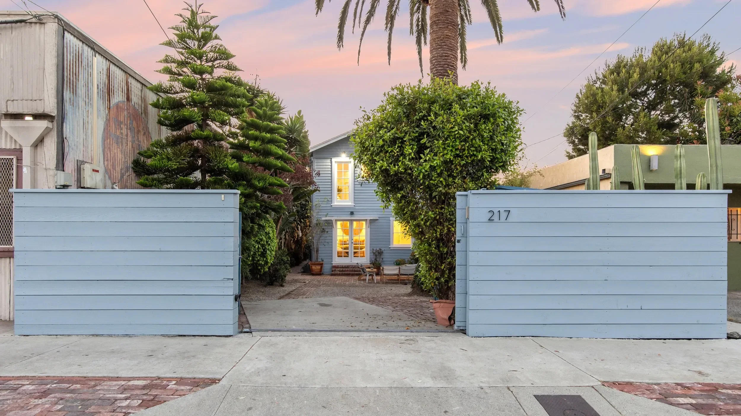A light blue wooden gate in front of a house with trees and shrubs, including a large palm tree, a pine tree, and a cactus, during sunset or dusk.