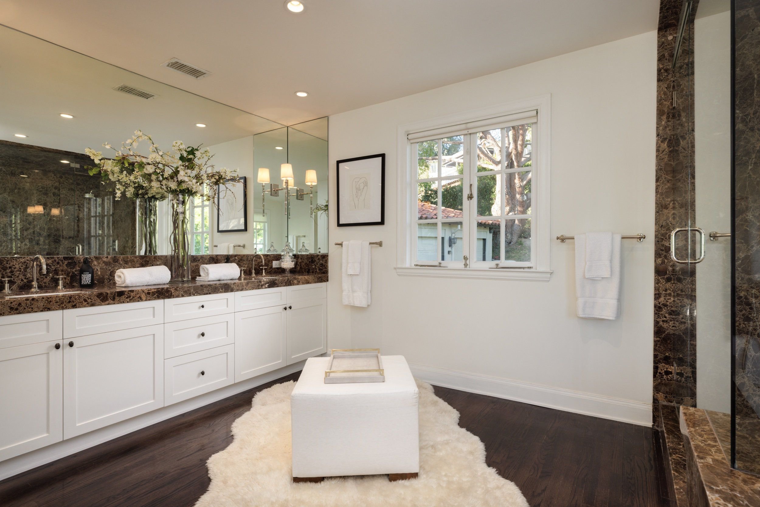 Bathroom with white cabinets, marble countertops, large mirror, wall-mounted lights, window showing outdoor trees, and a fluffy white rug.