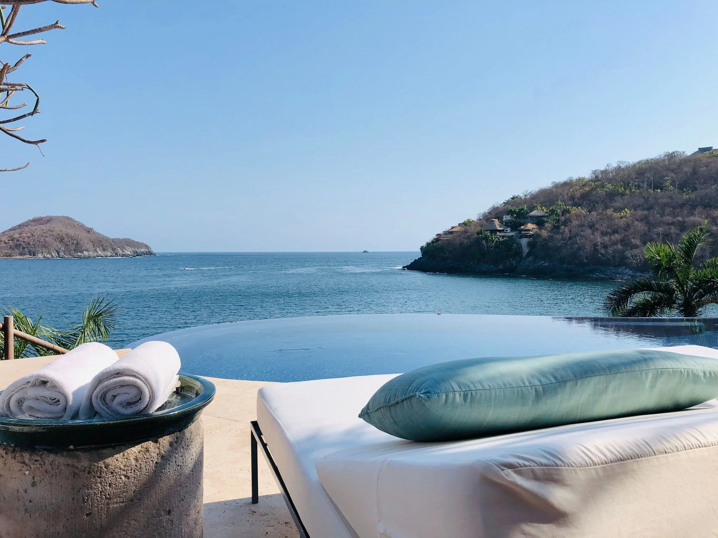 Beachside pool with rolled white towels on a black tray, lounge chair with a pillow, overlooking the ocean, hillside with houses, clear blue sky.