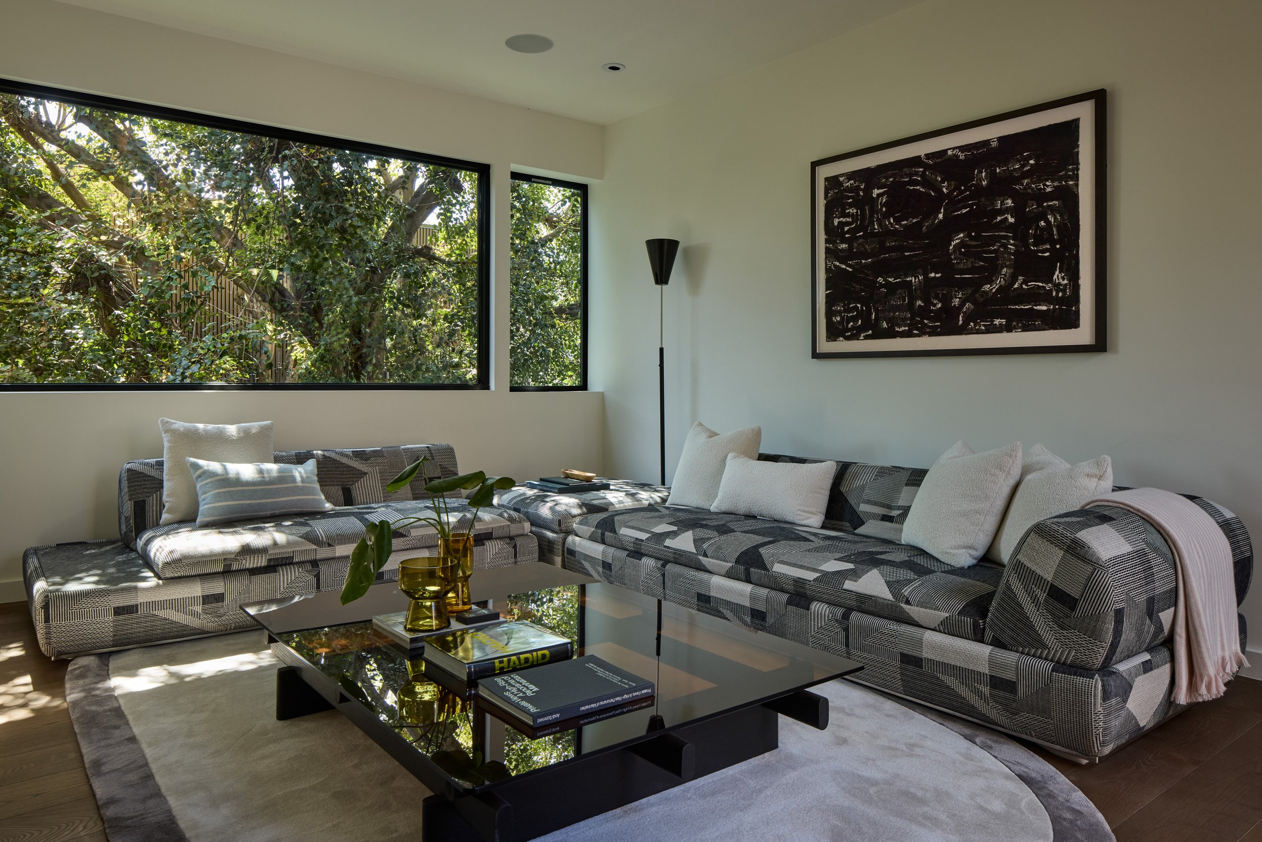 Modern living room with large windows overlooking trees, patterned sectional sofa with white and gray pillows, abstract black and white wall art, black floor lamp, glass coffee table with books and yellow vases, light-colored area rug, and throw blan