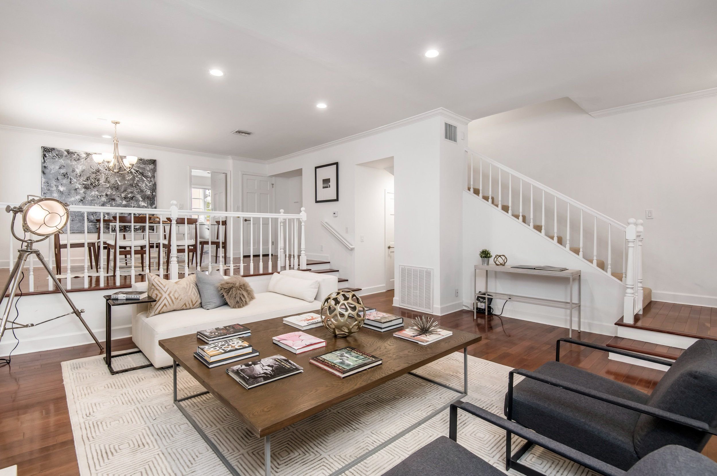 Living room with white walls, a white sofa with pillows, a wooden coffee table with magazines, black chairs, a large painting, and a staircase leading upstairs.
