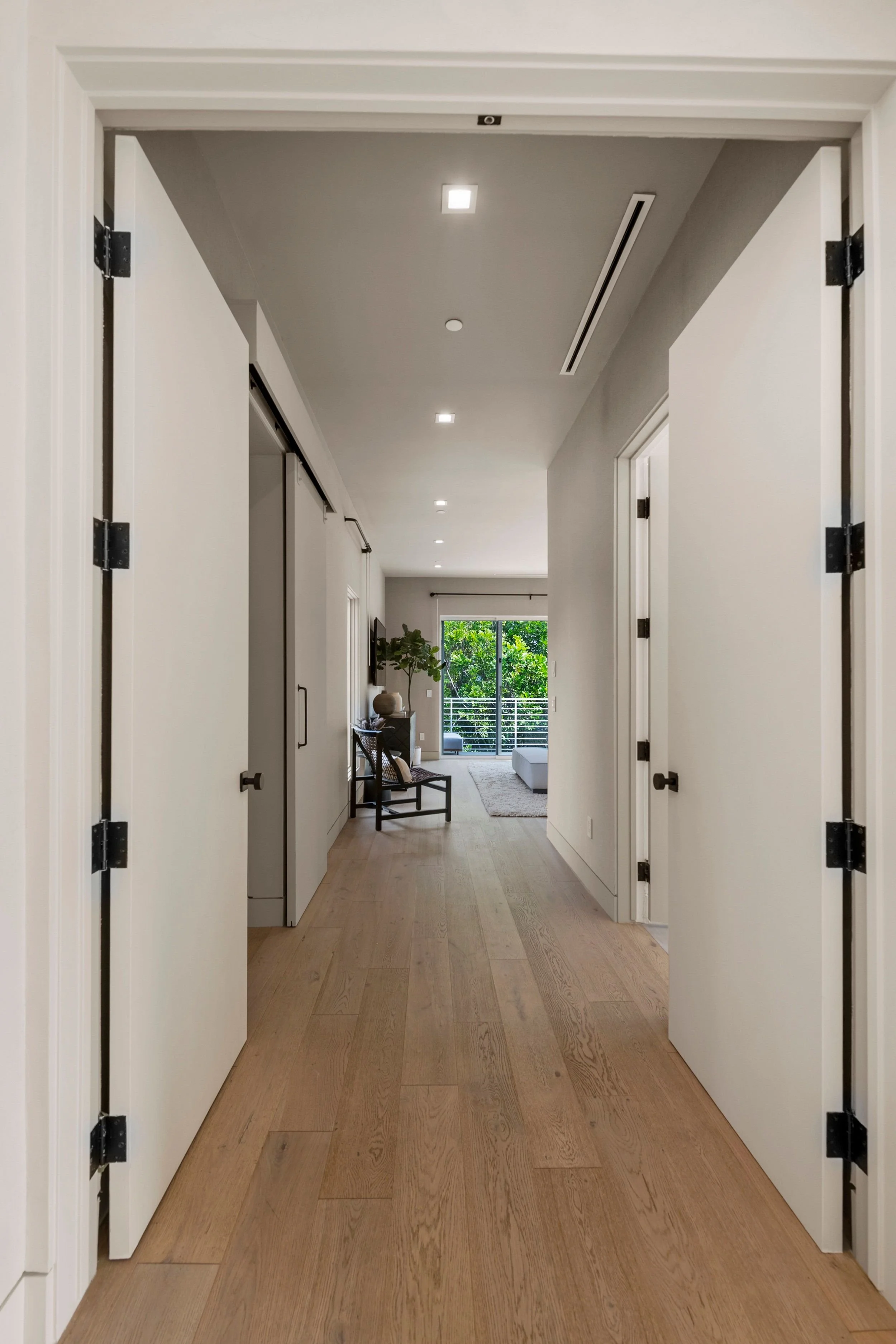 View of a modern hallway with wooden flooring, white walls, and a ceiling with recessed lighting. At the end of the hallway, there is a sliding glass door opening to a balcony with trees outside.