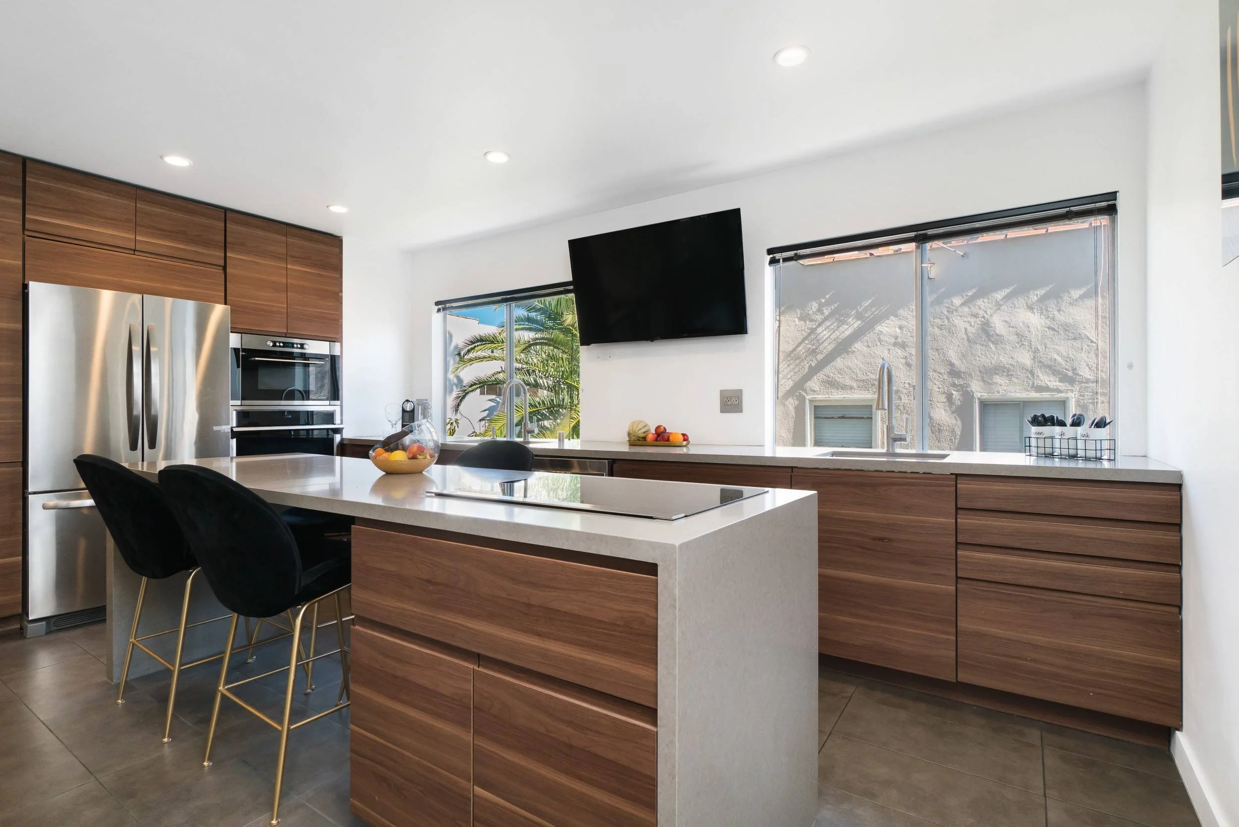 Modern kitchen with white walls, wooden cabinets, stainless steel refrigerator, built-in oven, black chairs at a white kitchen island, fruit bowl, large windows with blinds, and a wall-mounted TV.