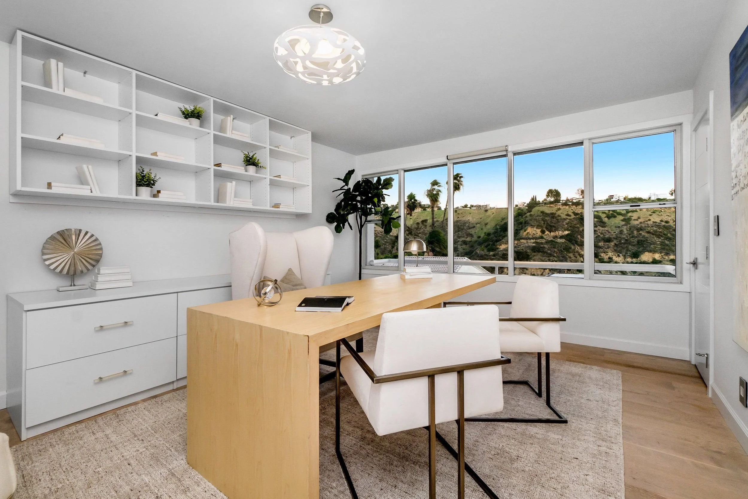 Modern office with a wooden desk, white chairs, a white bookshelf with plants, and large windows showing a hillside view of palm trees and houses.