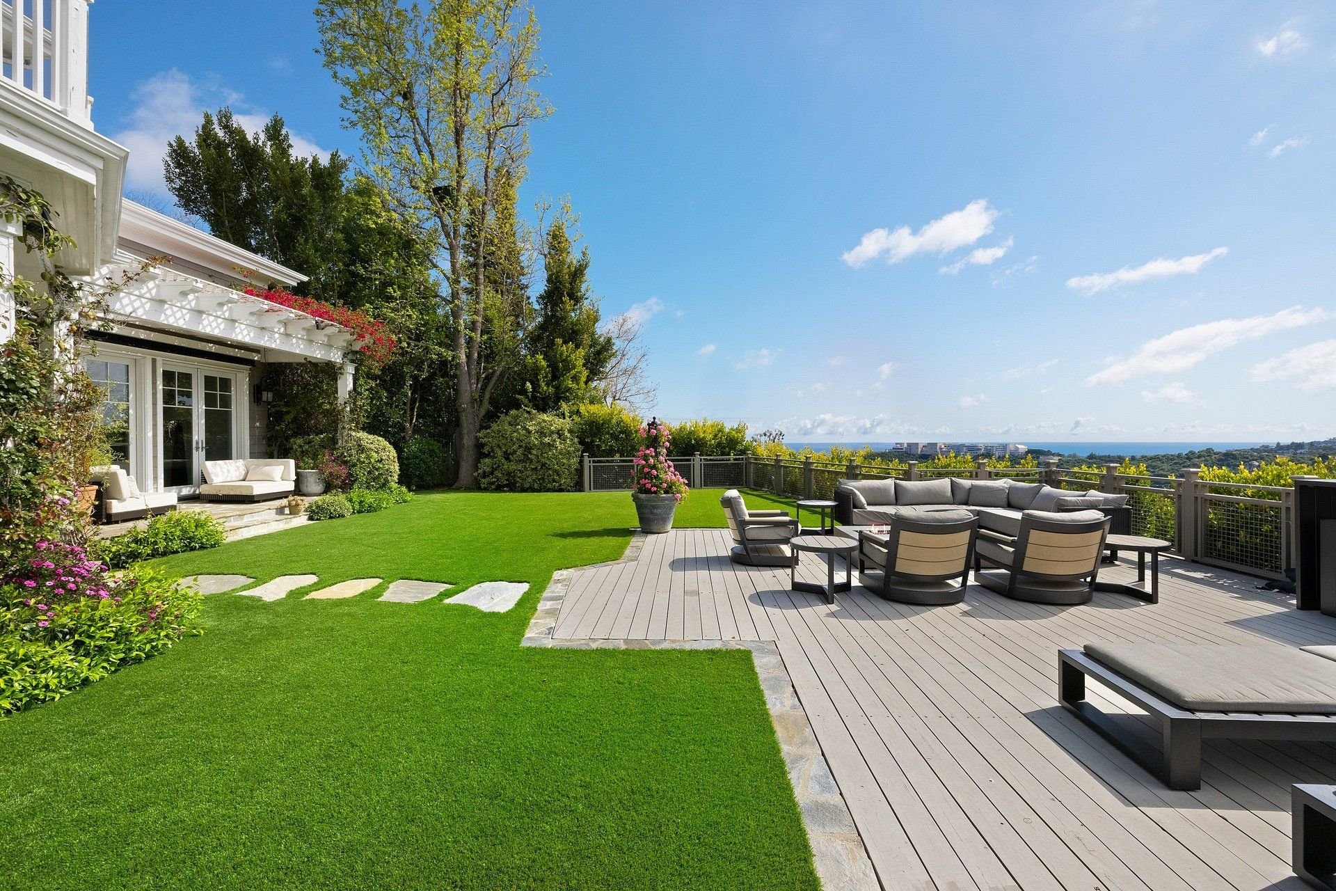 A backyard patio with outdoor furniture, a grassy lawn, flowering plants, and trees, with a view of the ocean and sky in the distance.