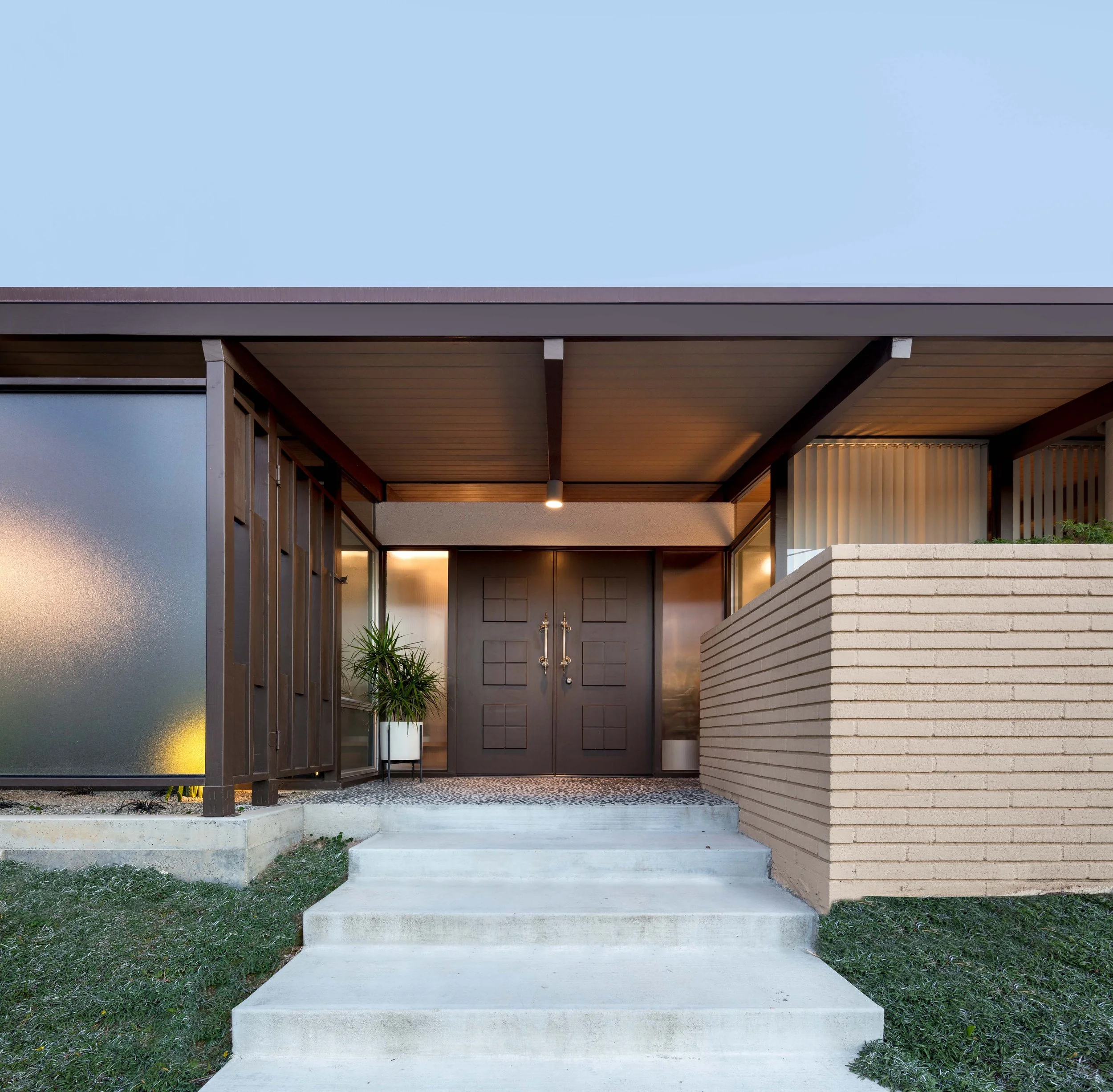 Modern house entrance with concrete steps, a porch with a wooden ceiling, and a brick wall on the right. A plant in a pot is near the door, and the sky is visible above.