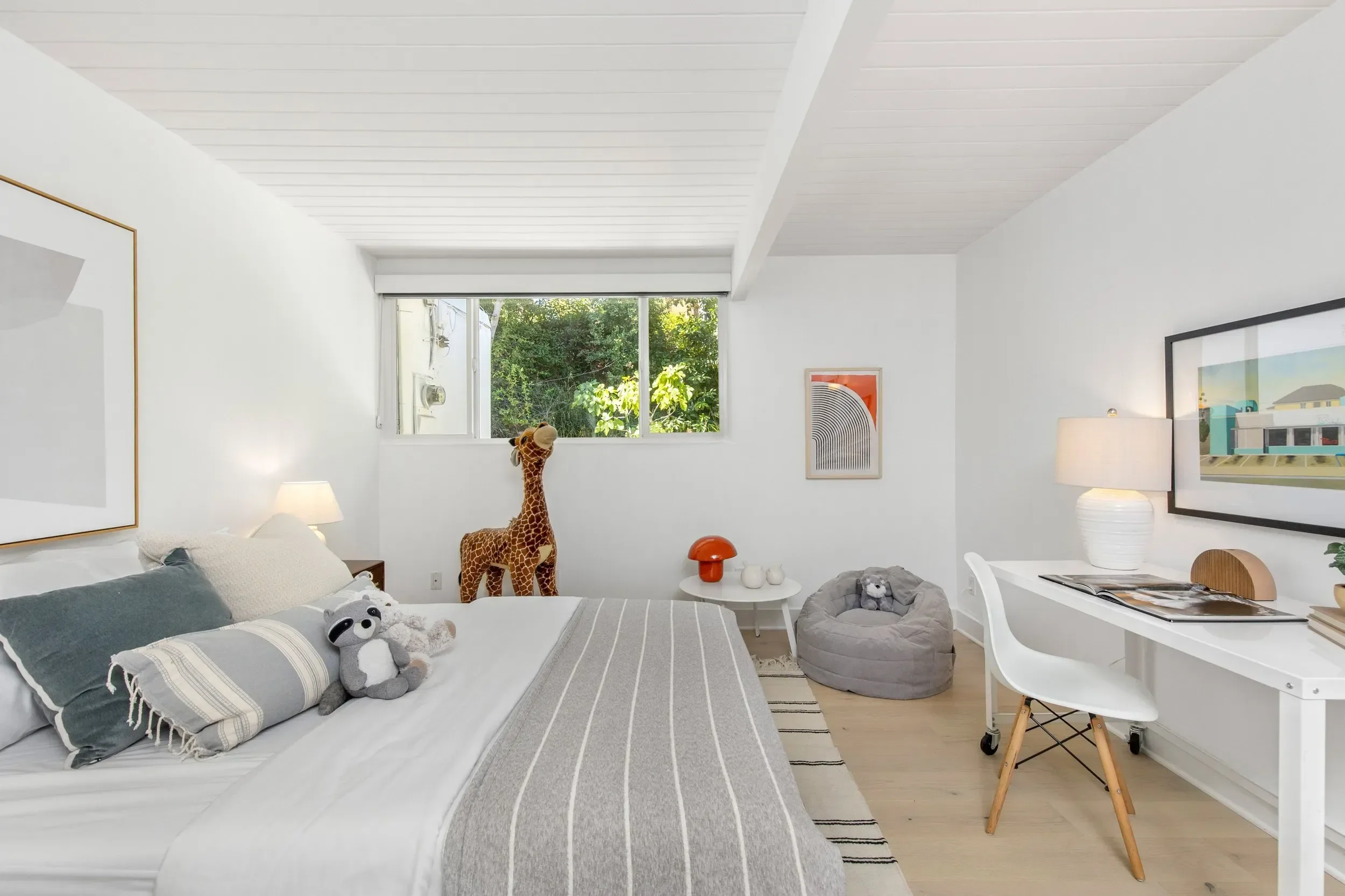 Bright, modern bedroom with white walls, large window inviting natural light, and various toys including a giraffe and panda on the bed and floor. Decor includes framed art and lamps, with a desk and books.