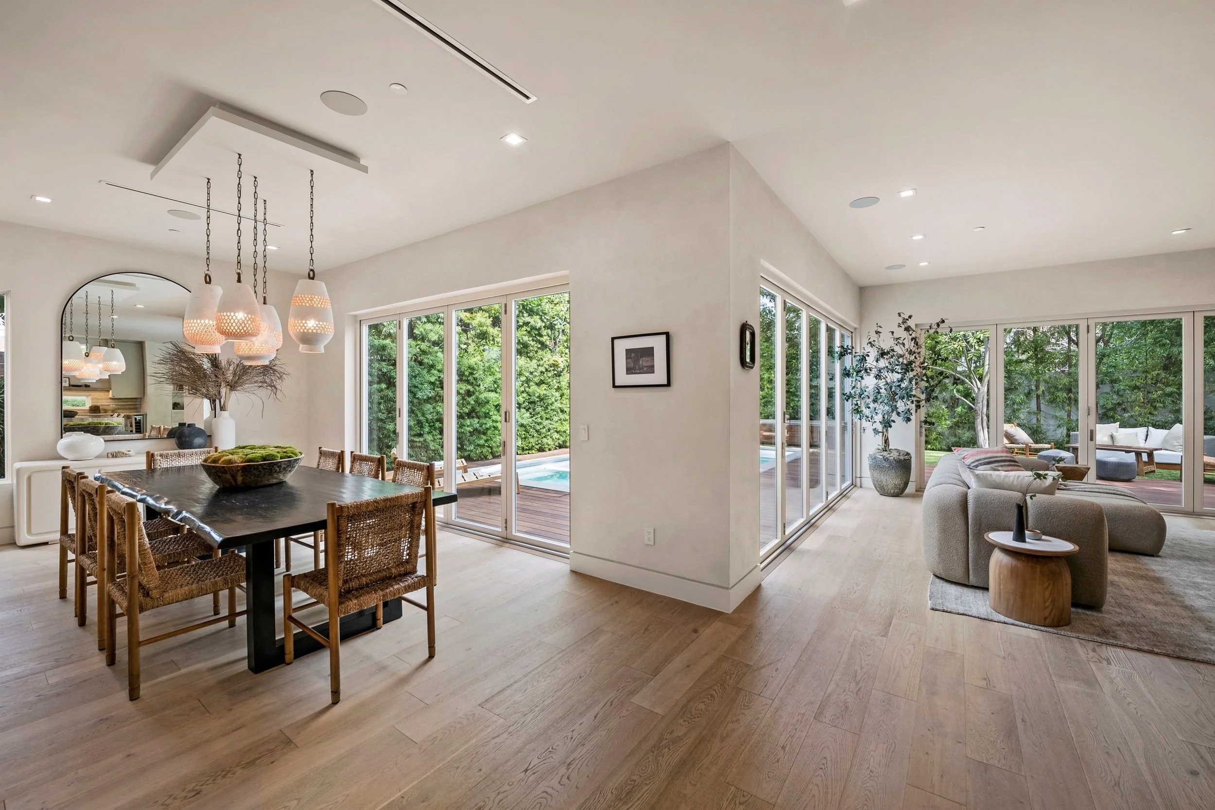 Interior of a modern home with an open floor plan, featuring a dining area with a rustic wooden table and wicker chairs, a living room with a gray sectional sofa, large sliding glass doors revealing a backyard with trees, and a white wall with framed