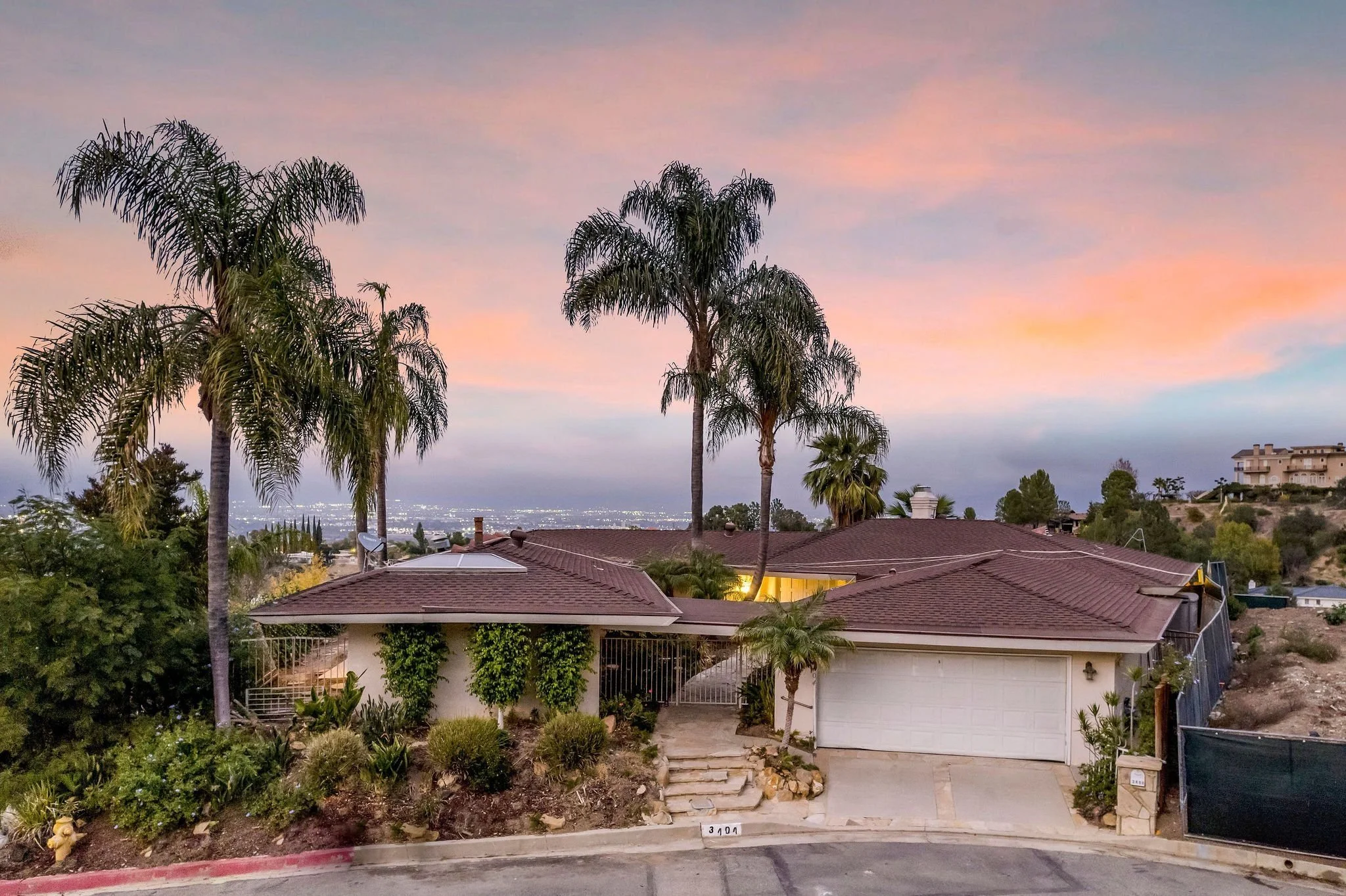 A suburban neighborhood with rooftops, palm trees, and houses under a pink and purple sunset sky.