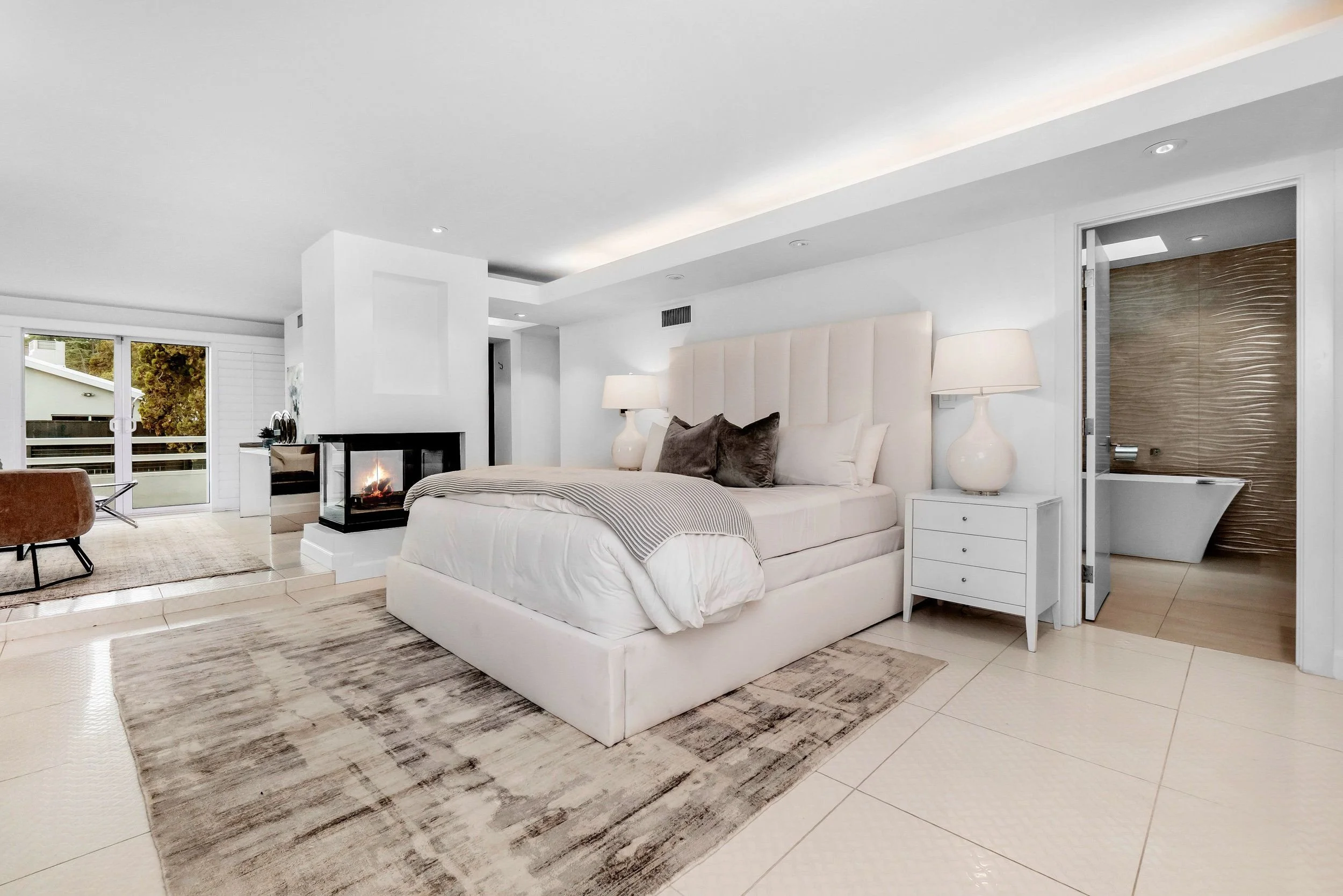 Modern bedroom with white walls, large bed with white headboard, bedside lamps, a fireplace, sliding glass doors leading to a balcony, and a bathroom with a bathtub and textured wall.