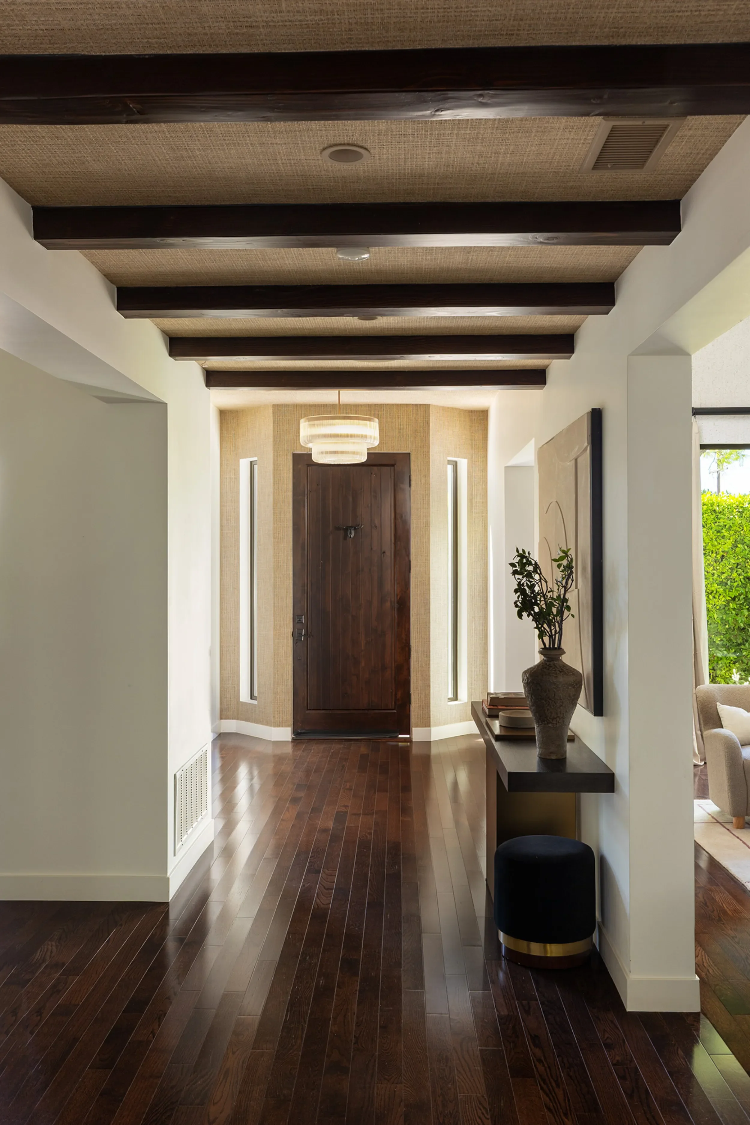 An entryway with dark wooden flooring, white walls, and a wooden door at the end. There is a round light fixture hanging from the ceiling, flanked by narrow windows on each side. A small console table with a tall vase and some books is on the right, 