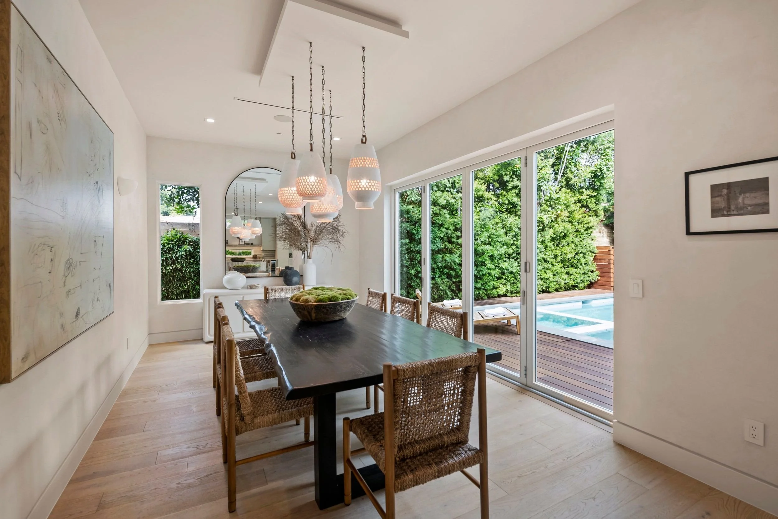 A modern dining room with a dark wooden table surrounded by woven rattan chairs, pendant lights hanging above, and large sliding glass doors leading to a backyard with trees and a small pool.