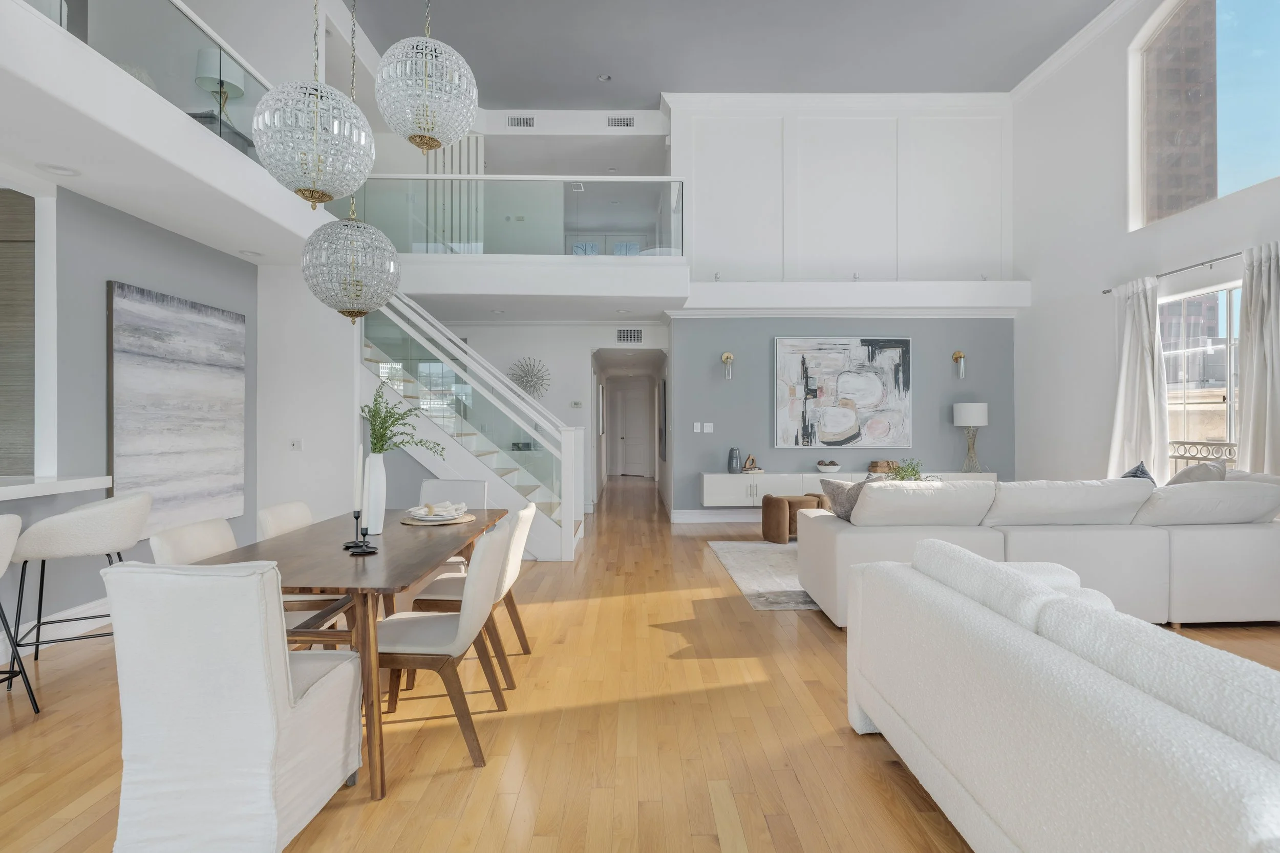 A spacious modern living room with light wood flooring, white and gray walls, and large windows with white curtains. There is a white couch, a piece of abstract art on the wall, a wooden dining table with white chairs, and decorative lighting fixture