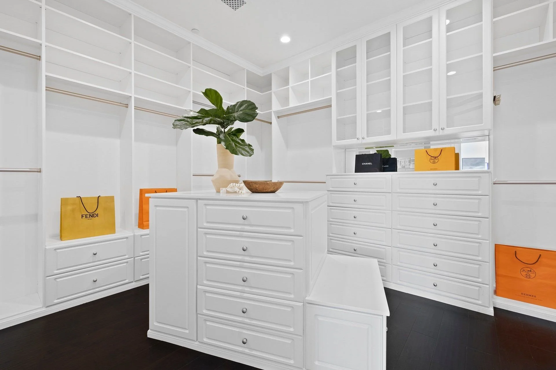 A walk-in closet with white cabinetry and black flooring, containing luxury shopping bags from brands such as Fendi, Chanel, Hermès, and Louis Vuitton, along with a large green plant in a beige vase and decorative objects on the countertop.
