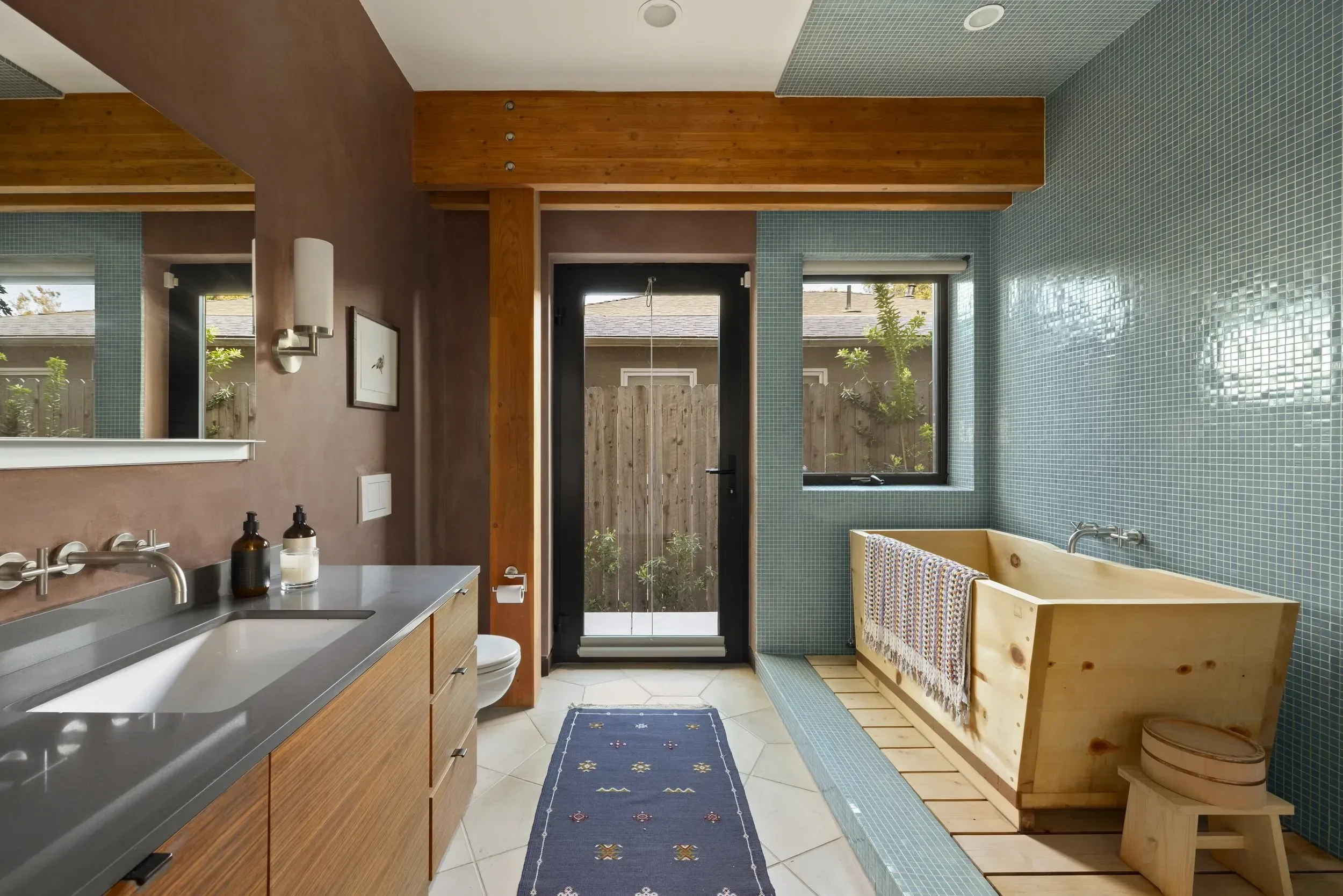 A modern bathroom with a wooden vanity, a gray countertop, and a sink. There is a wall-mounted light fixture and a framed picture on the brown-painted wall. The bathroom has windows and a door leading outside, with greenery visible. There is a blue r