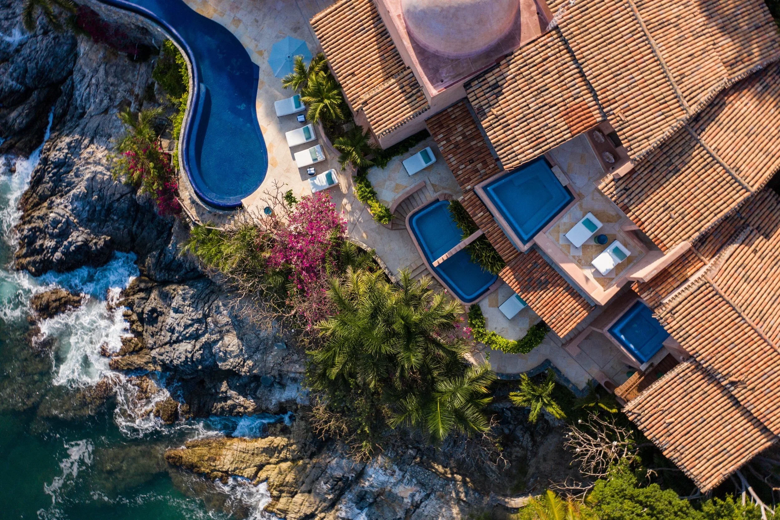 Aerial view of a seaside resort with swimming pools, palm trees, and tiled roof buildings near rocky coast and ocean.