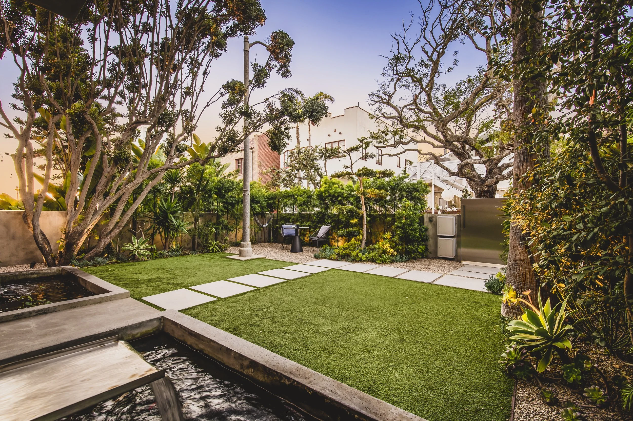 A backyard garden during sunset with trees, a lawn, a stone pathway, and outdoor furniture, including two chairs and a table.