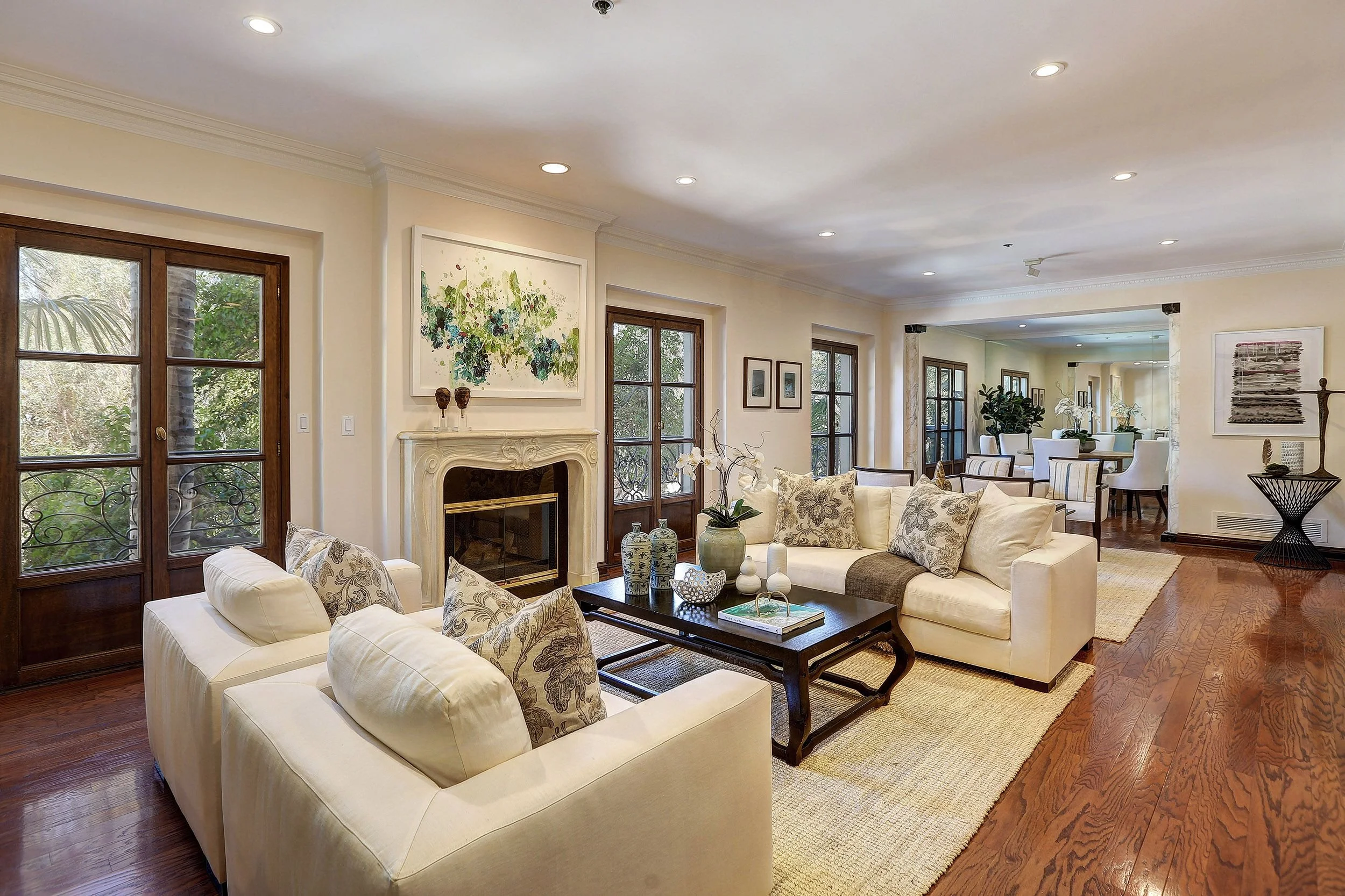 A spacious living room with cream-colored walls, hardwood floors, and multiple large windows with wooden frames. The room features white sofas with patterned cushions, a dark wooden coffee table with decorative vases and books, and artwork on the wal