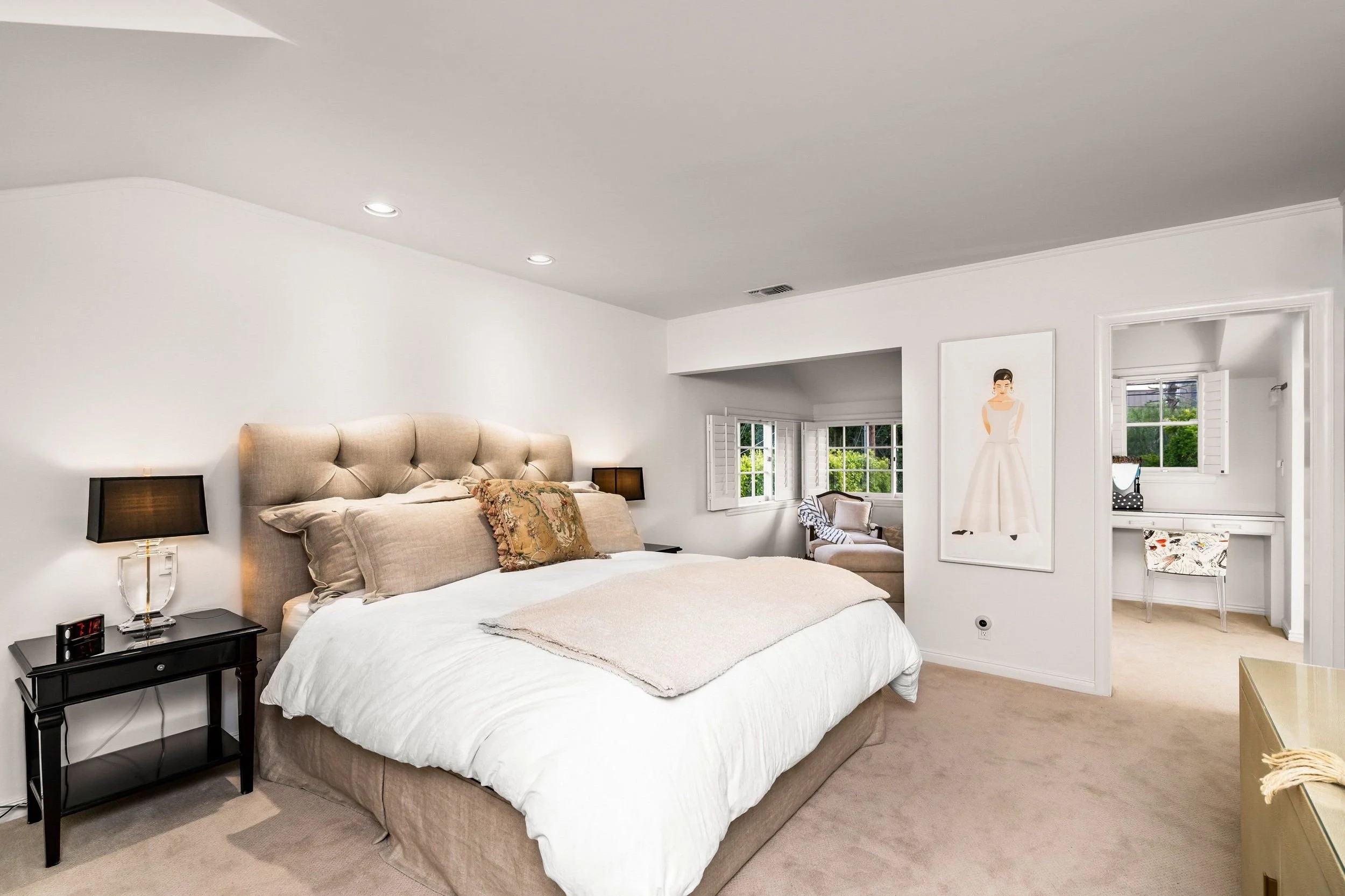 A cozy bedroom with a large bed featuring beige upholstery, white bedding, and decorative pillows. On each side of the bed are black nightstands with matching lamps. The room has white walls, carpeted floors, and multiple windows with white shutters,
