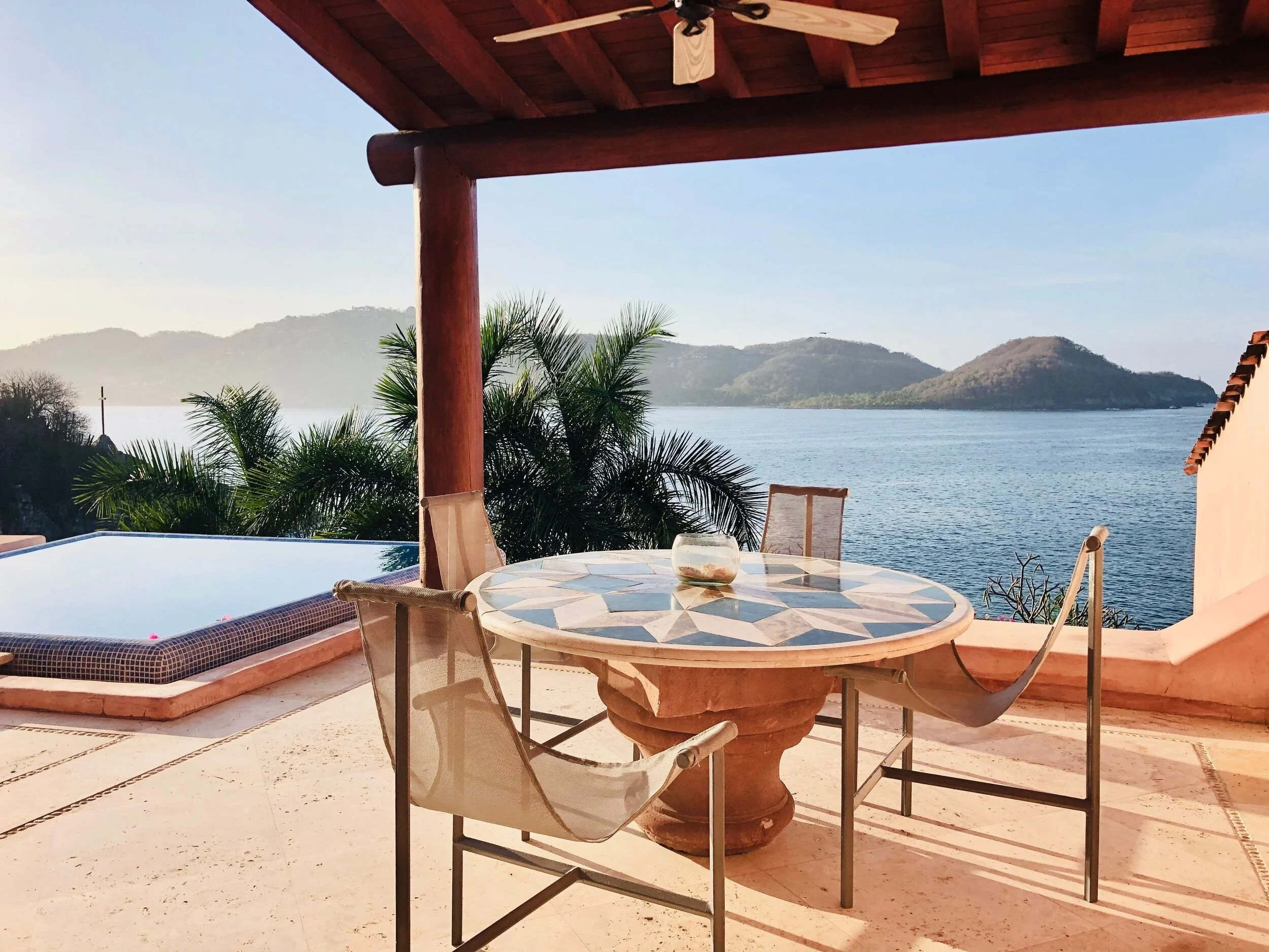 A patio with a round mosaic table, surrounded by chairs, overlooking a body of water with hills in the distance. There is an infinity pool to the left, a wooden pergola overhead with a ceiling fan, and lush palm trees nearby.