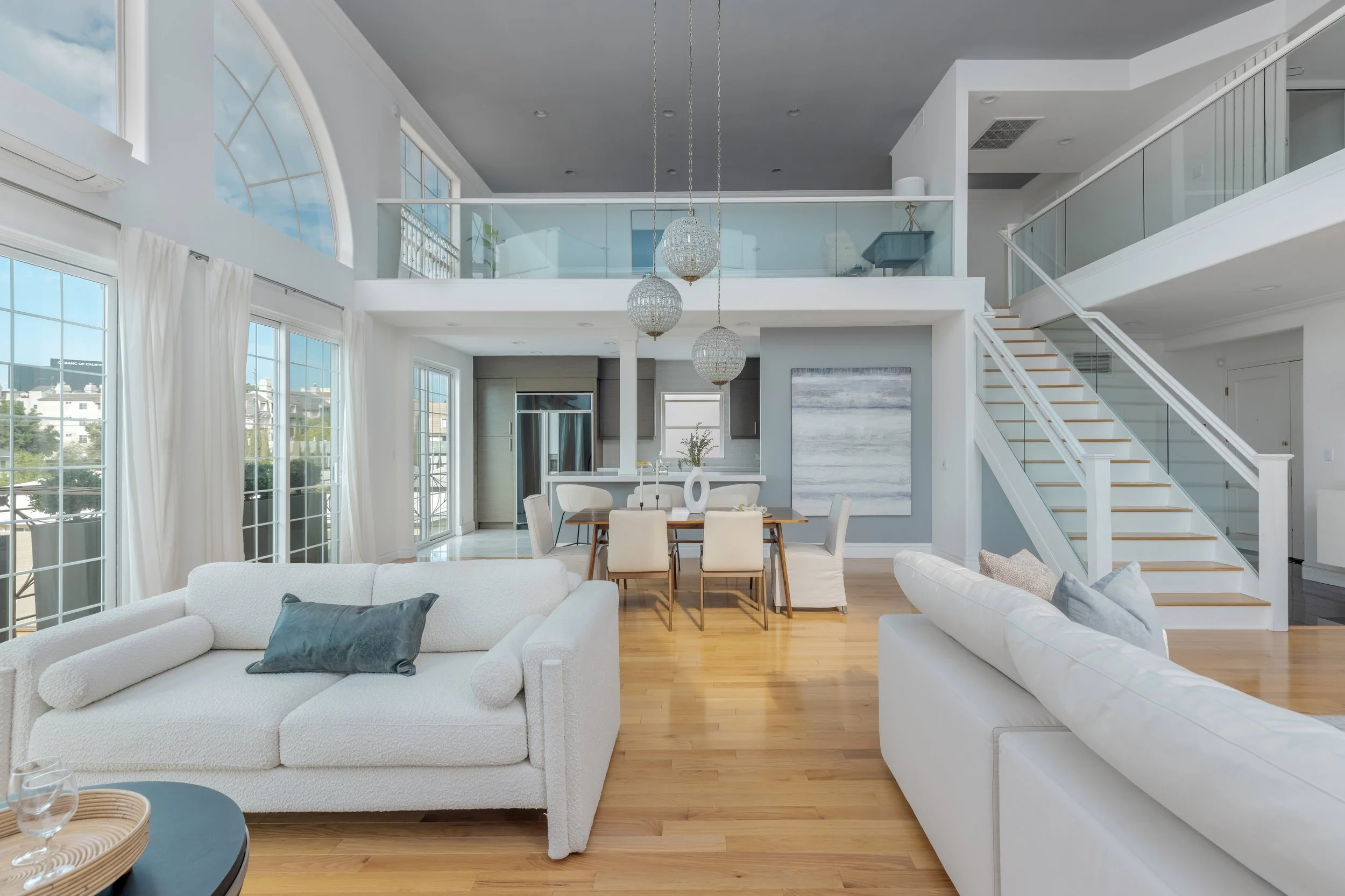 Bright, modern living room with white sofas, wooden flooring, large windows with white curtains, a dining area with six chairs, and a staircase leading to upper floor. Three hanging spherical light fixtures and a painting on the wall are visible.
