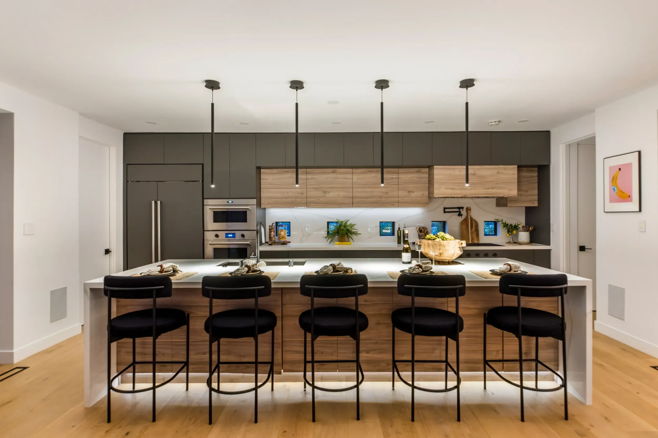 Modern kitchen with black and wood cabinetry, a large island with five black barstools, pendant lighting, and small framed artwork on the walls.