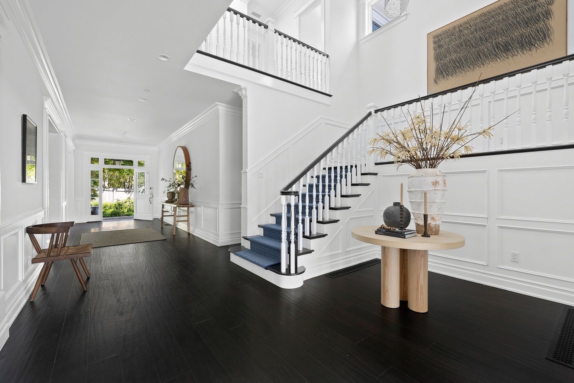 Entrance foyer with staircase, dark wood flooring, white walls, and modern decor including a large vase with dried plants and a round table with decorative items.
