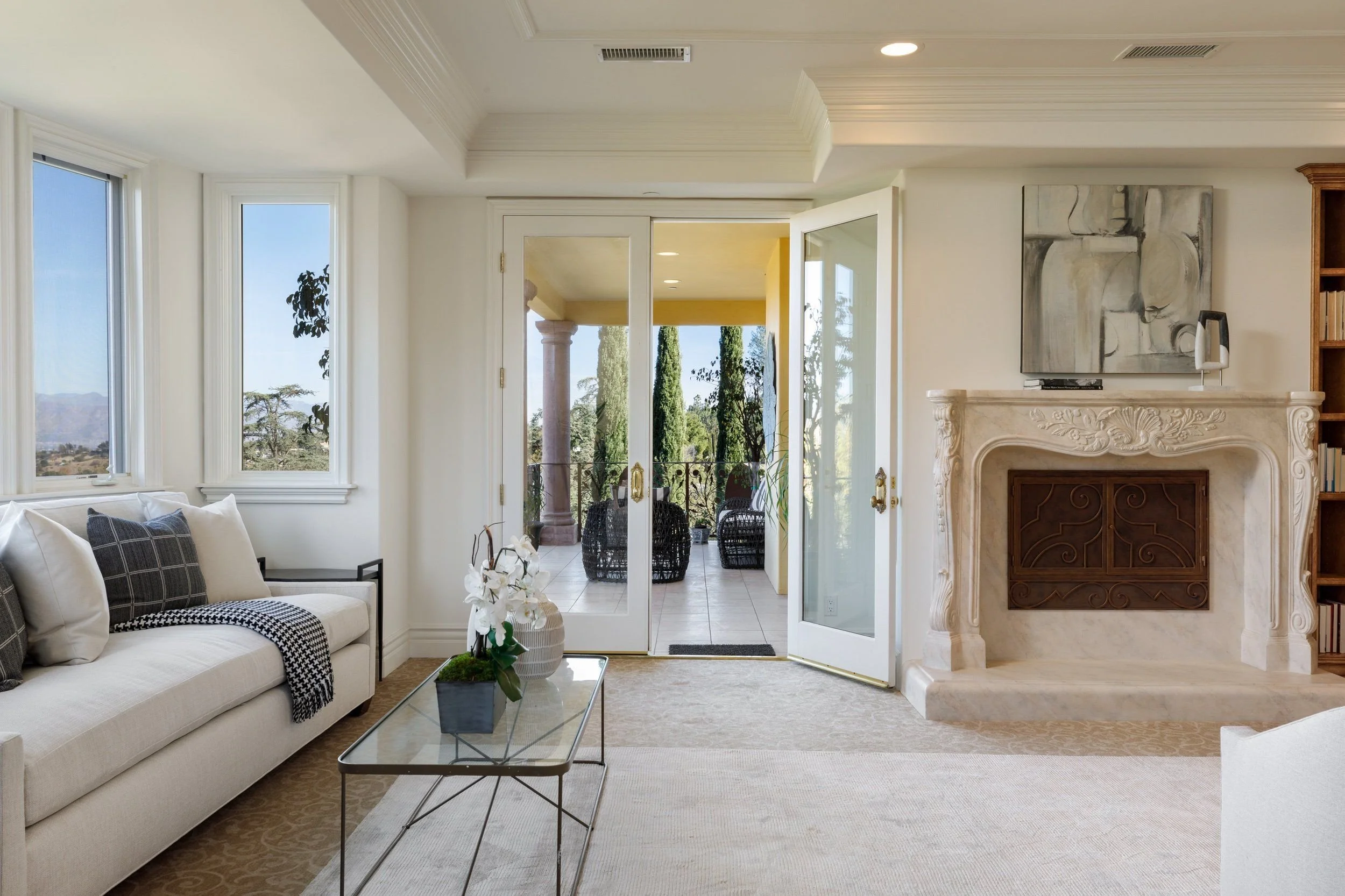 A bright living room with white walls, large windows, a cream sofa with black and white pillows, a glass coffee table with a plant, and a fireplace with abstract black and white artwork above it. French doors lead to an outdoor patio with wicker chai