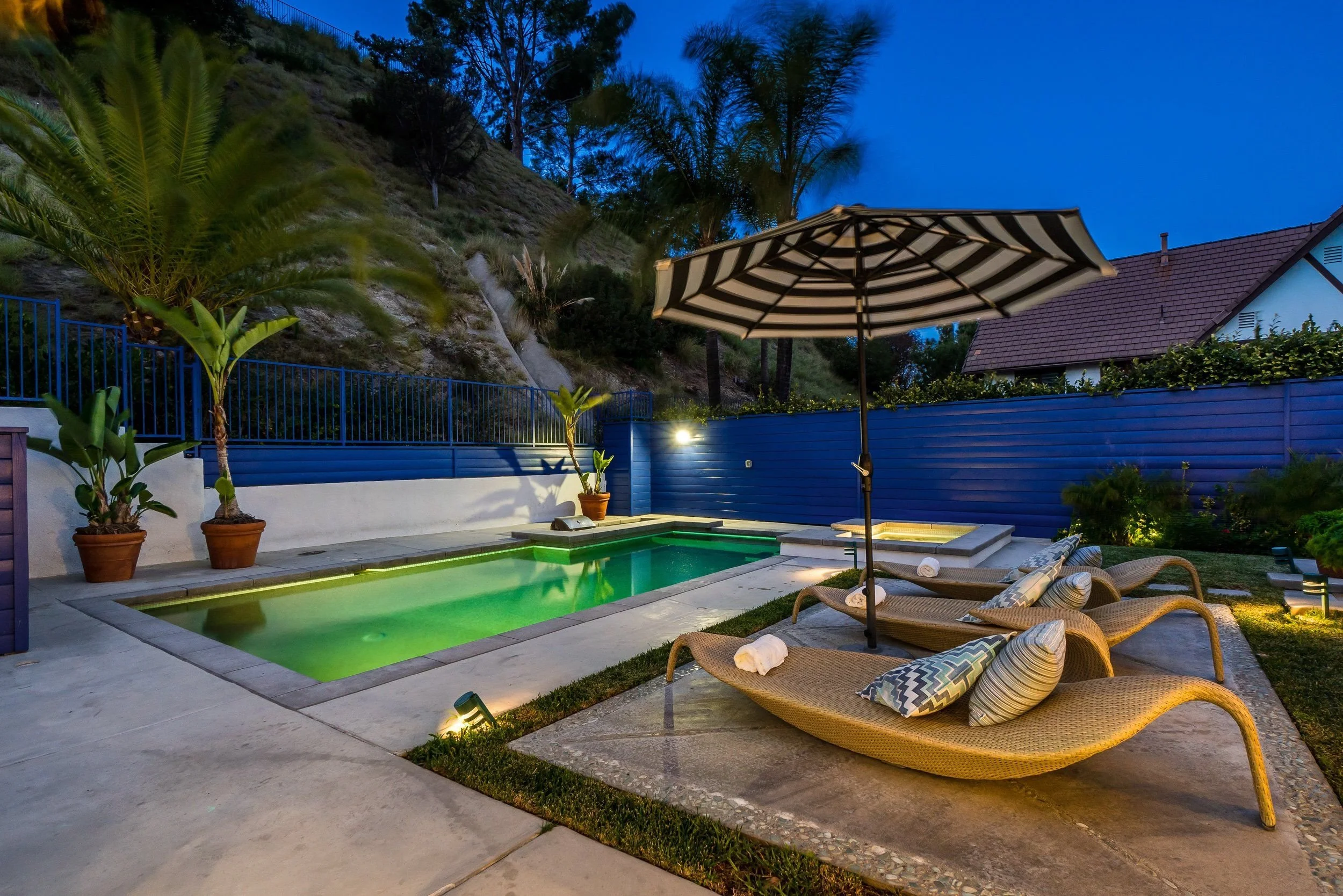 Backyard outdoor swimming pool with lounge chairs, pillows, a large umbrella, potted plants, and a blue fence. Hills and trees are visible in the background during dusk.