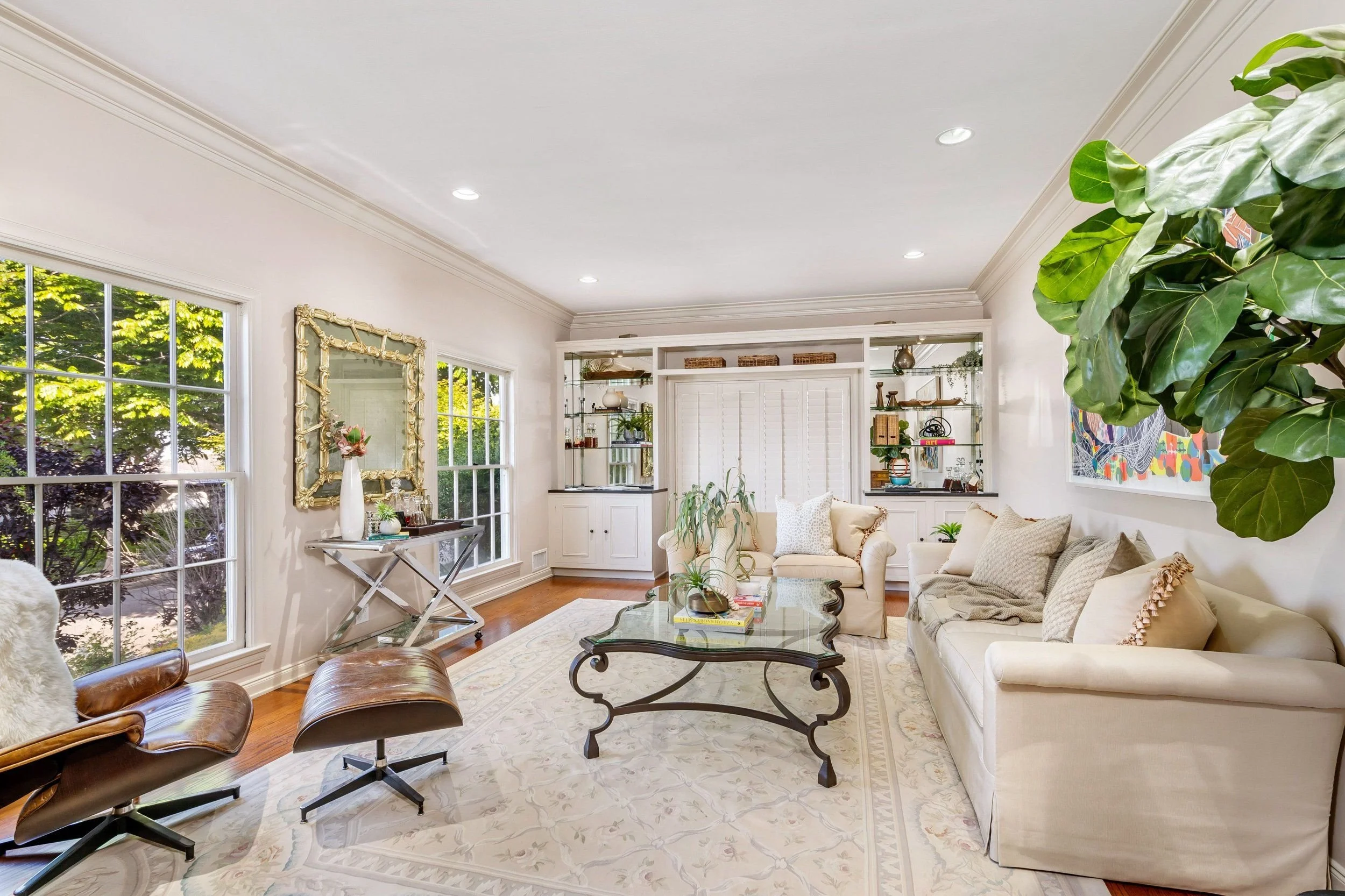 Bright living room with large windows, cream-colored sofas with pillows, a glass coffee table, and indoor plants, with a view of greenery outside.