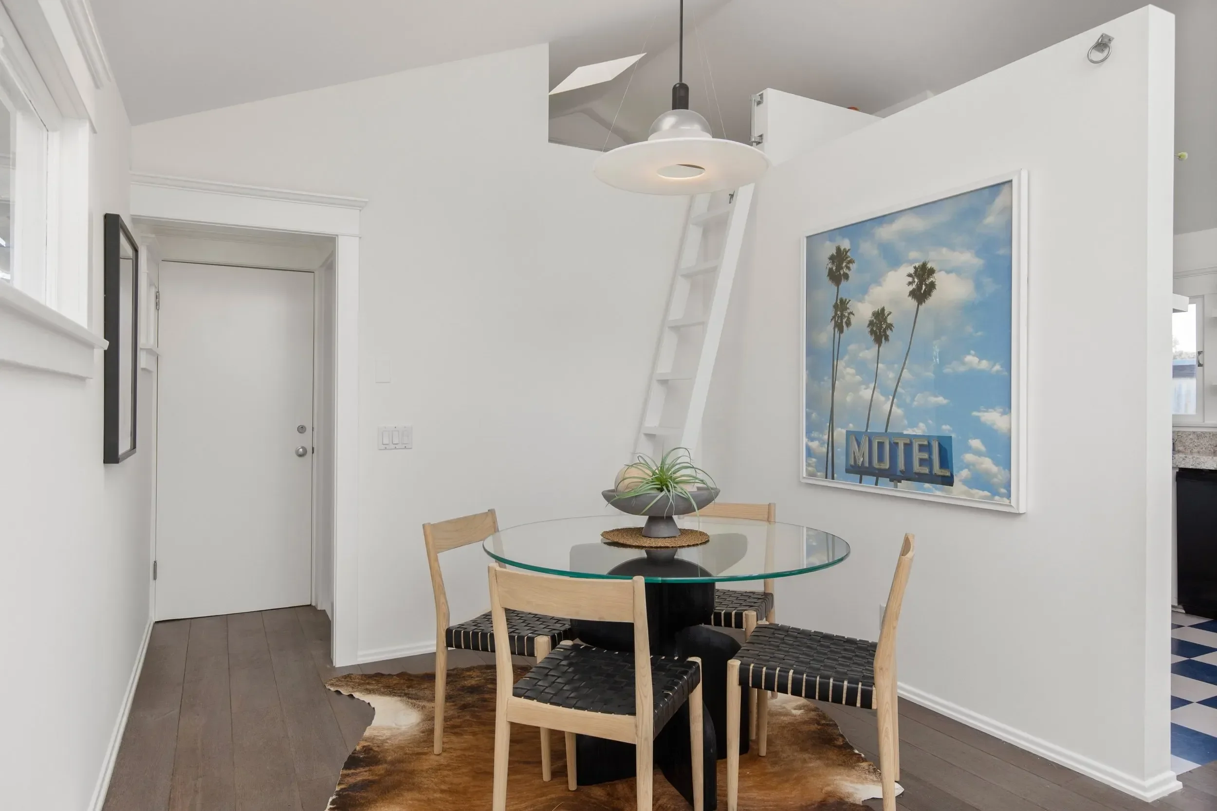 A dining area with a glass round table, four wooden chairs with woven black seats, a cowhide rug, a white wall with a large picture of a Motel sign and palm trees, a wall-mounted mirror, and a white ladder leaning against the wall.