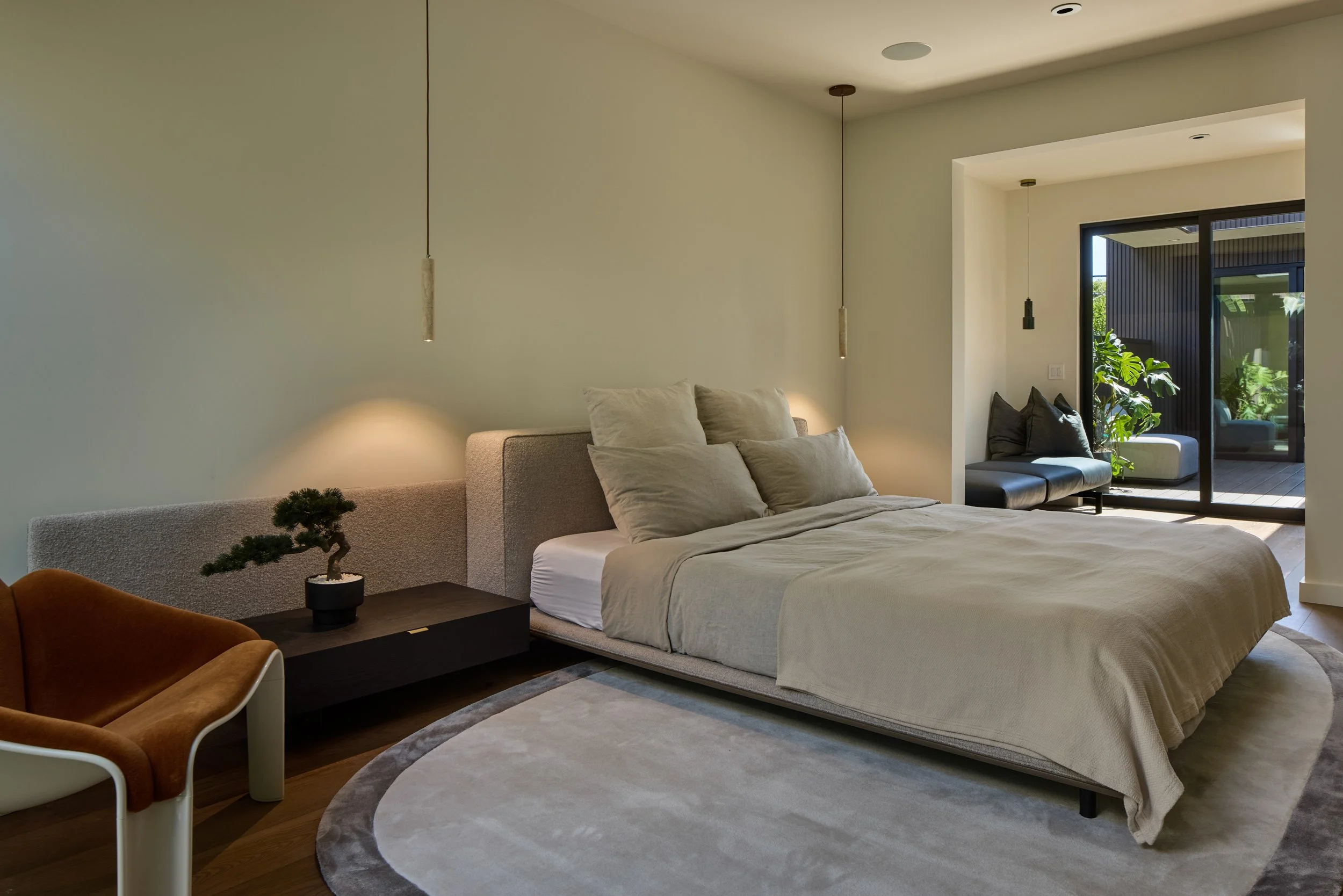 Modern bedroom with a large bed, beige bedding, and multiple pillows, a small side table with a potted plant, and a sliding glass door leading to a balcony with green plants outside.