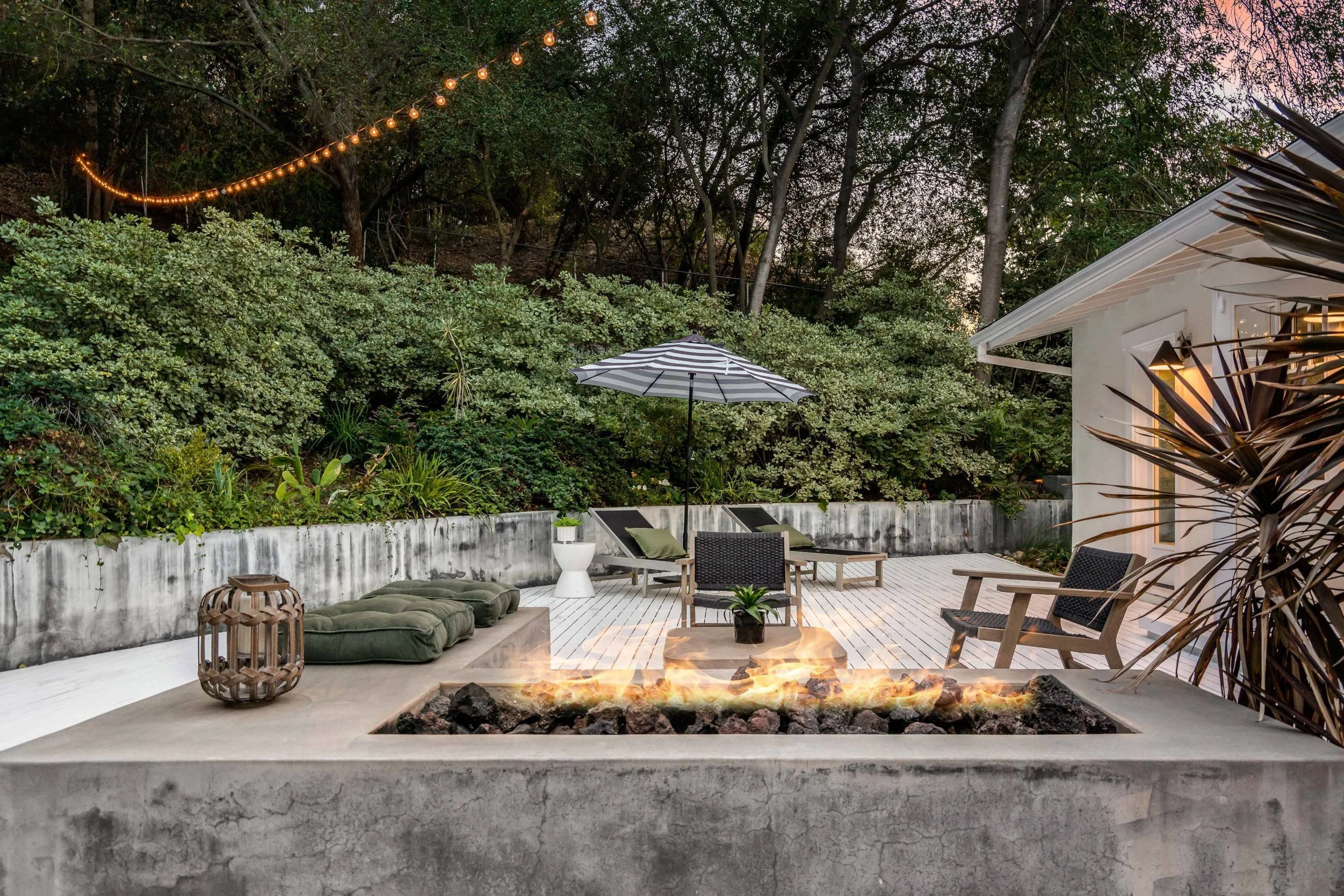 Modern outdoor patio with a built-in fire pit, seating, a striped patio umbrella, and lush greenery at dusk, with string lights overhead.