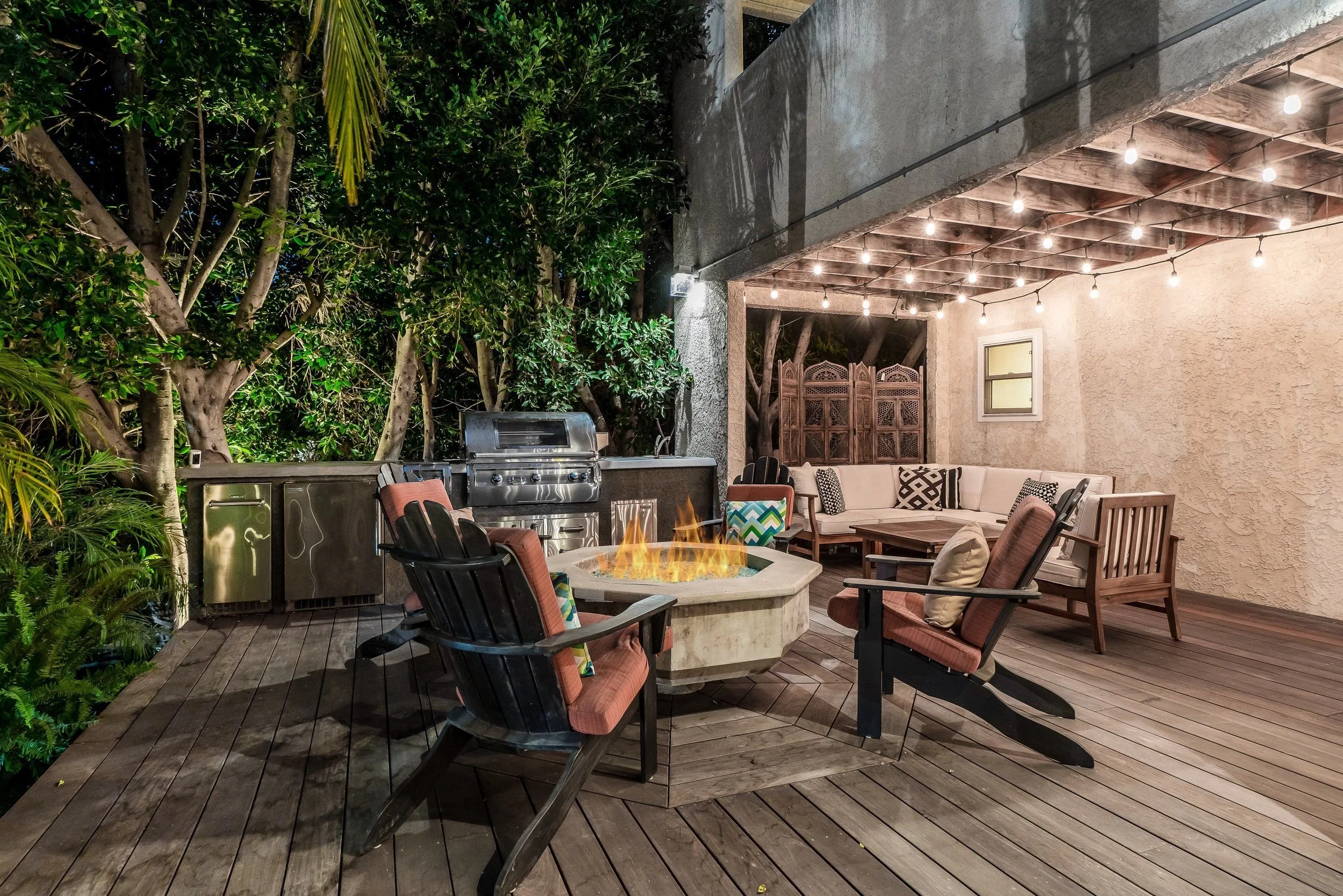 An outdoor patio at night with string lights overhead, a built-in fire pit with flames, cushioned chairs, a wooden outdoor sofa, a grill, and surrounded by greenery and trees.