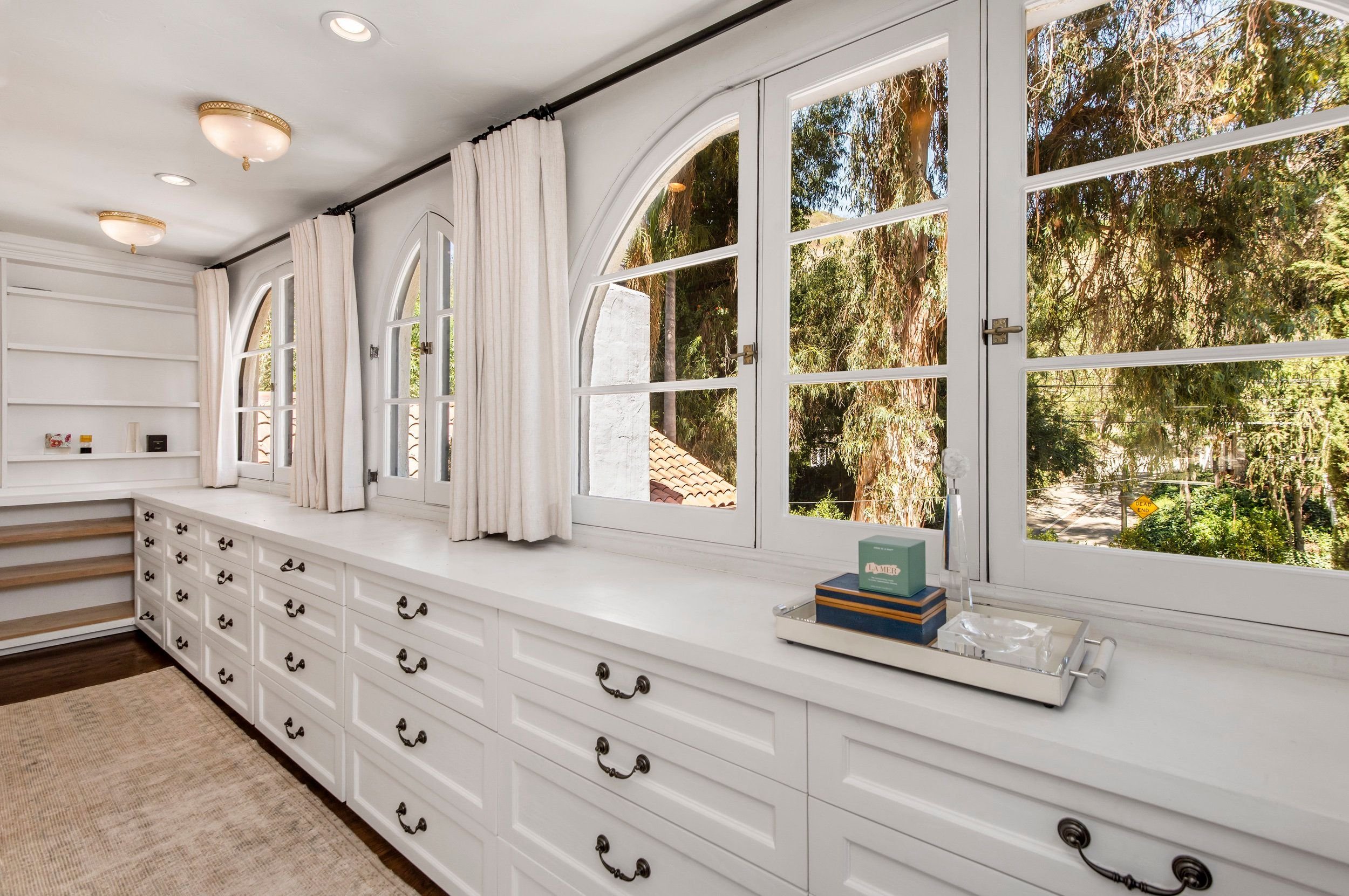 White built-in cabinet with multiple drawers and black handles, topped by a white countertop, beneath large windows with white curtains, overlooking trees outside.
