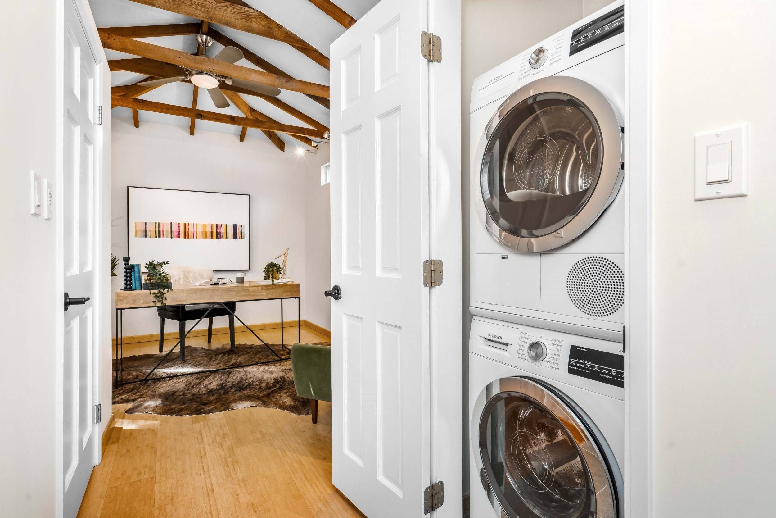 A view into a laundry room with a stacked washer and dryer on the right, a white wall with a light switch, and a home office area with a desk, chair, and artwork on the wall in the background.